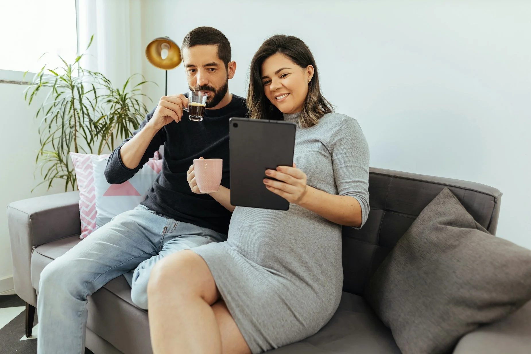 A pregnant woman and a man sitting on a sofa in a living room, looking at a tablet together. The woman is holding a pink mug, and the man is drinking coffee from a clear glass.