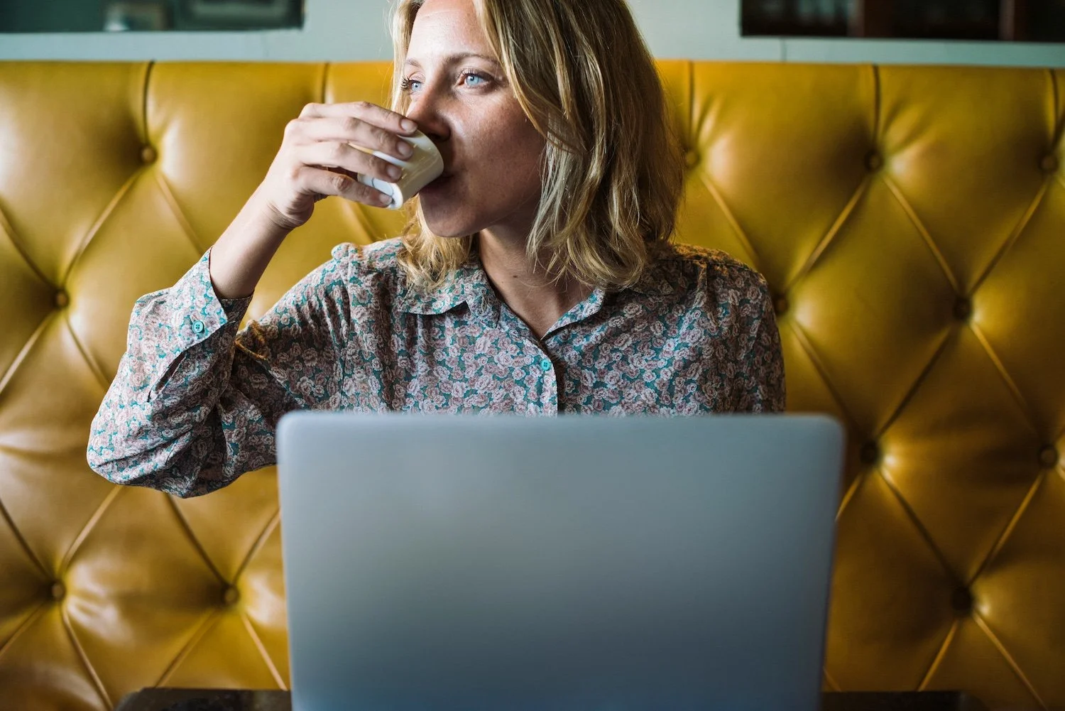 A woman with blonde hair and blue eyes drinking coffee while using a laptop at a NYC cafe.