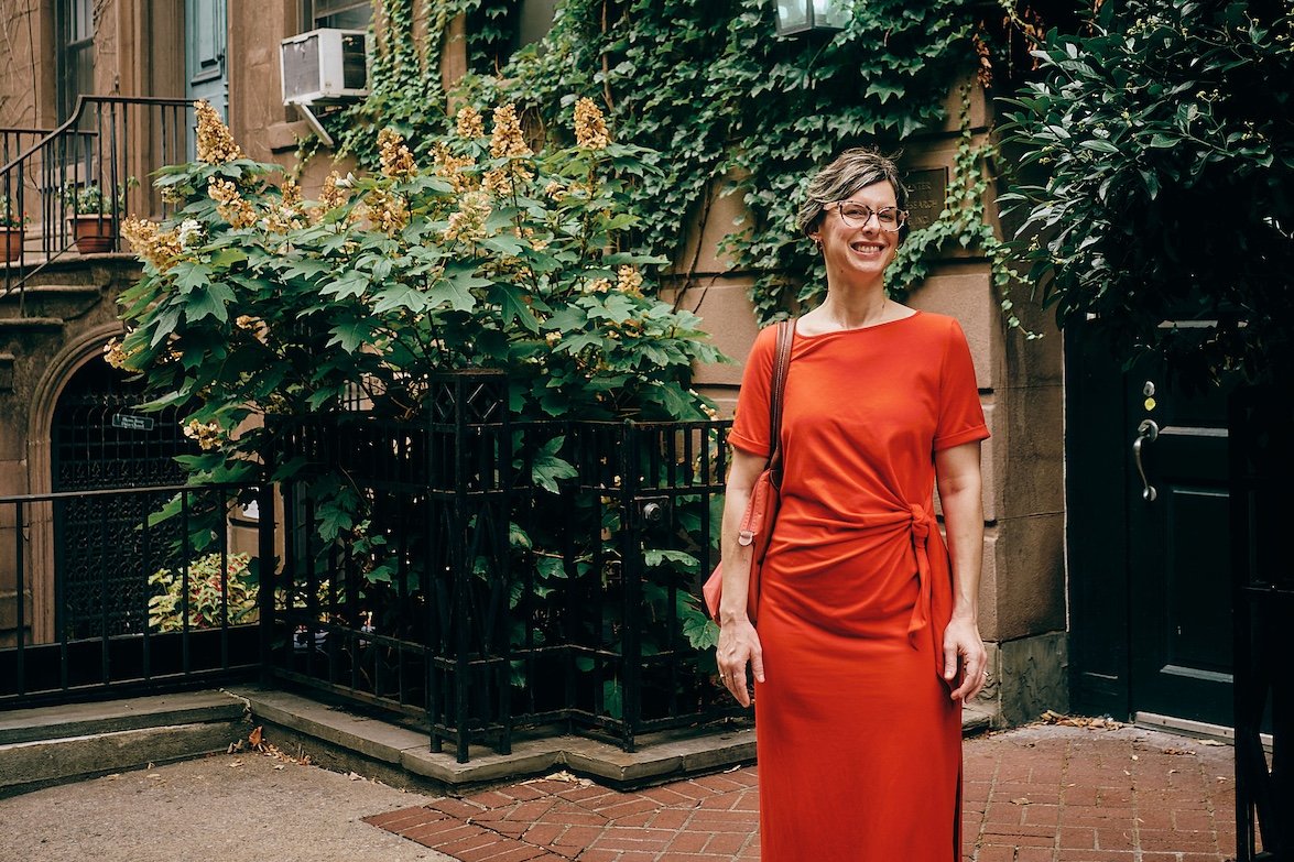 Photo of Barbara Isenberg, owner of Fiscal New Yorker. She's smiling with glasses and short dark hair, standing outdoors in front of leafy plants and a building in New York City. She is wearing an orange dress and carrying a shoulder bag.