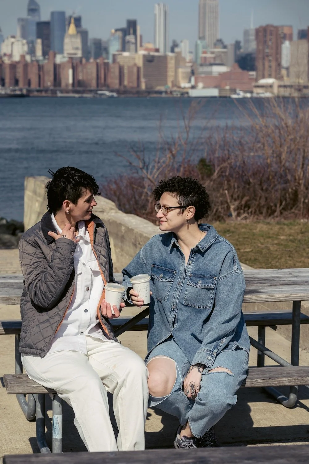 Two women sitting on a park bench by a body of water, holding coffee cups, engaged in conversation with the New York City skyline in the background.