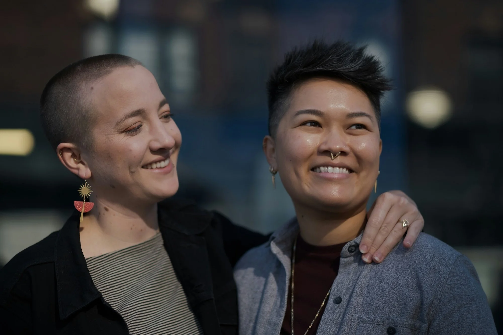 Two women smiling and looking happy, one with a shaved head and wearing jewelry, the other with short hair and a nose ring, embracing outdoors with a blurred urban background.