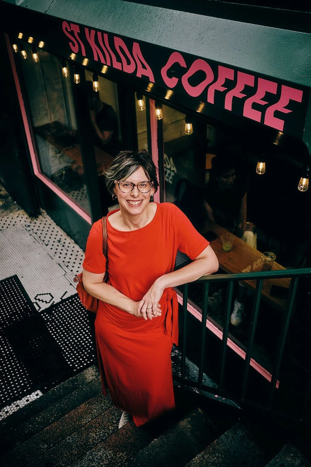 Barbara Isenberg, owner of Fiscal New Yorker, smiling in a red dress, standing on a staircase next to a coffee shop, looking up at the camera, with the shop's pink sign reading 'St Kilda Coffee' visible above.