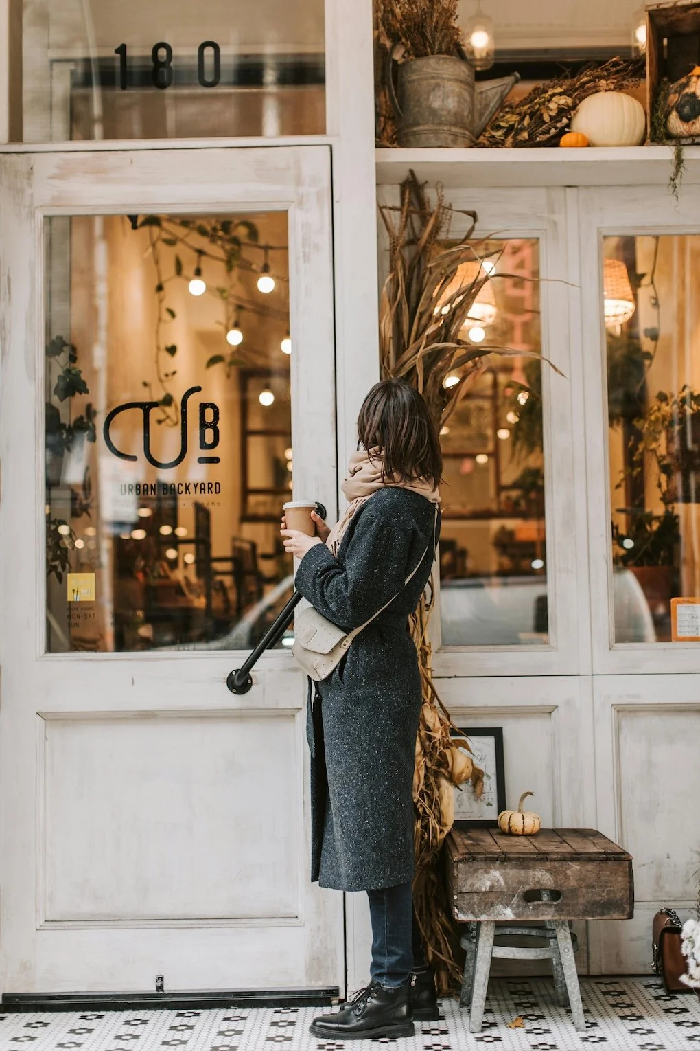 A woman in a dark coat and boots stands outside a cozy New York City cafe, holding a coffee cup, with autumnal decor including pumpkins and dried plants visible around her and through the window of the cafe.