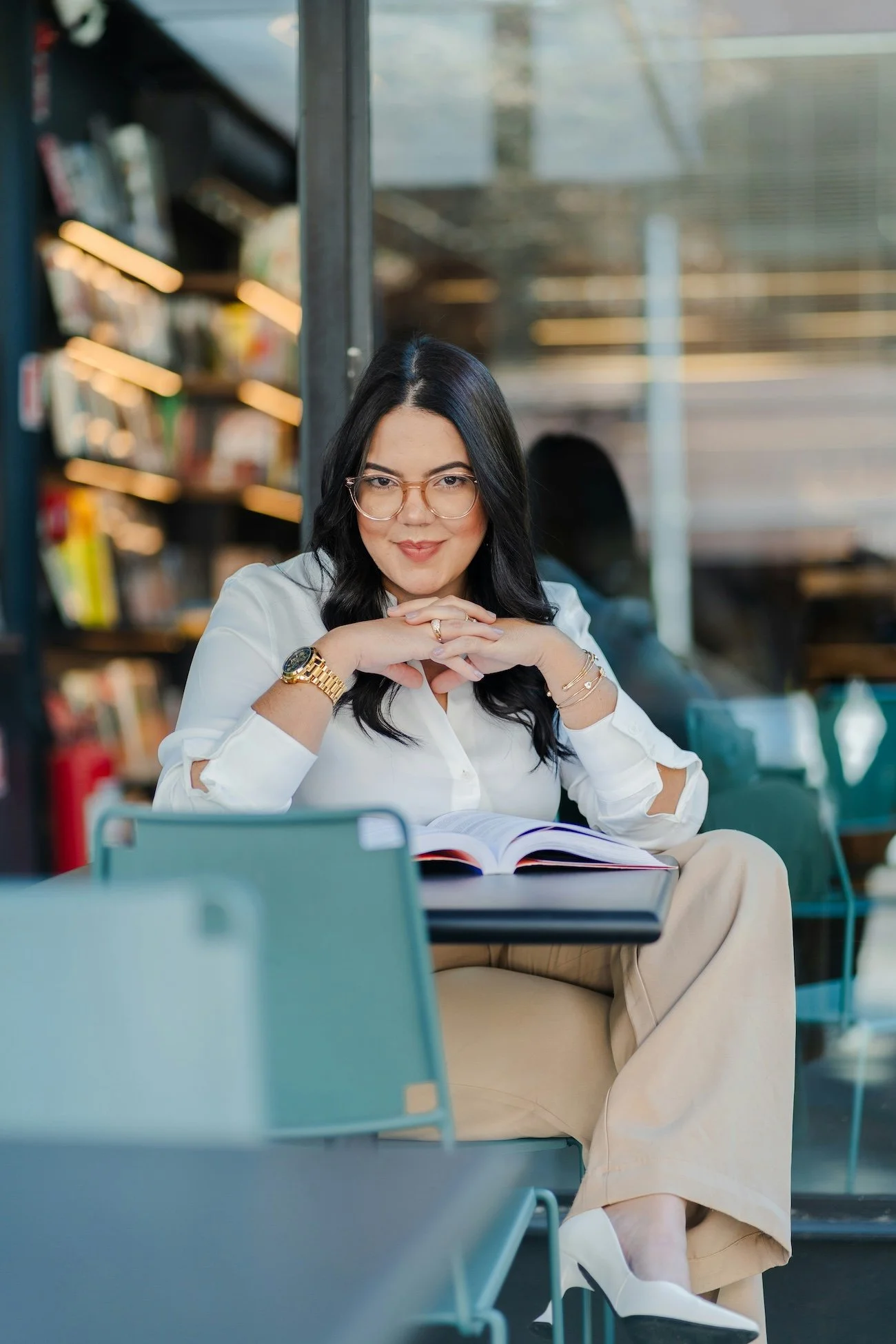 A woman with black hair, glasses, and wearing a white shirt sitting at an outdoor table in New York City with an open book in front of her. She is smiling and looking at the camera with her hands clasped under her chin.
