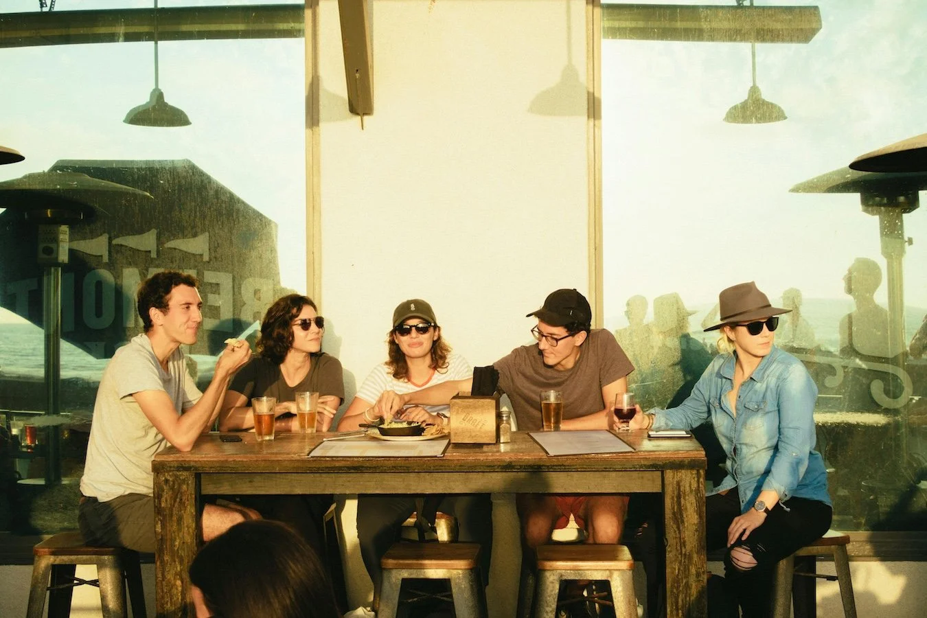 Five friends sitting at a wooden table in a New York City restaurant, enjoying drinks and conversation, with large glass windows showing a view outside, during sunset.