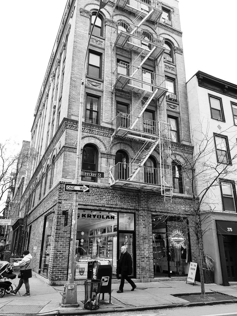 A black and white photo of a multi-story brick building in New York City with fire escape stairs on the front, a storefront at the ground level, street signs, and pedestrians on the sidewalk.