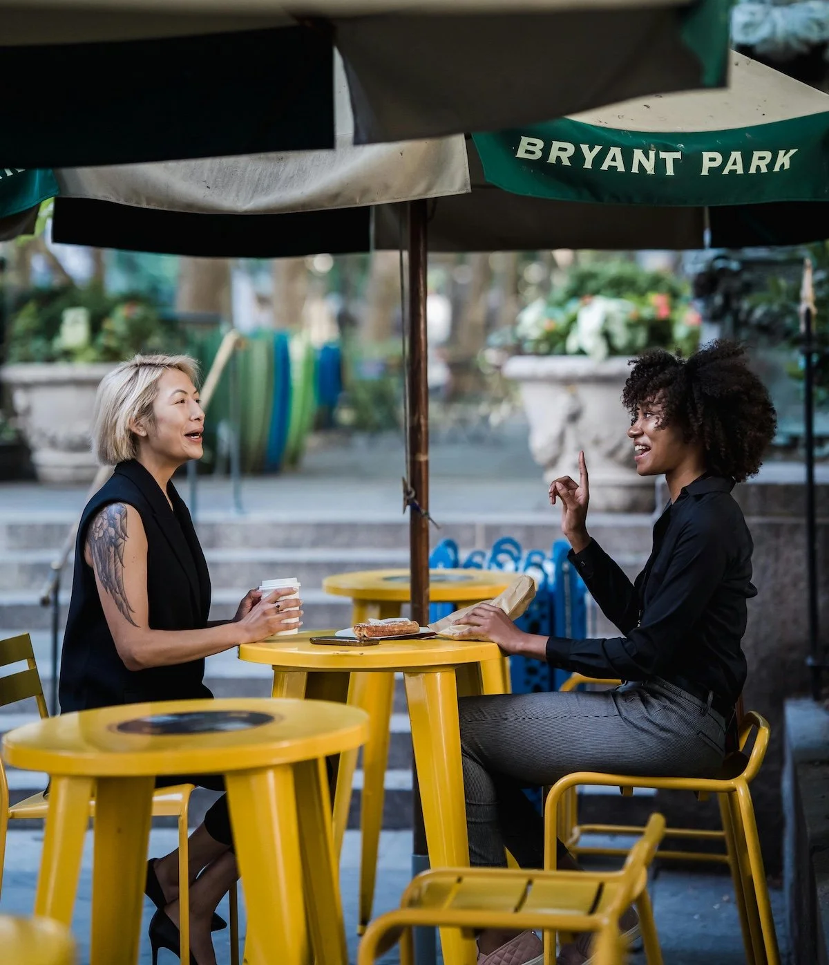Two women sitting at yellow outdoor cafe tables under a Bryant Park umbrella, engaging in conversation. One woman has blonde hair, tattoos on her arm, and is holding a coffee cup; the other woman has curly dark hair and is gesturing with her finger.