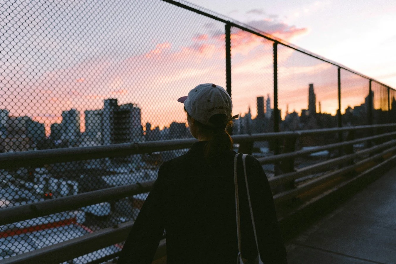 A woman wearing a baseball cap and black jacket walking on a bridge at sunset with the New York City skyline in the background and a chain-link fence along the bridge.