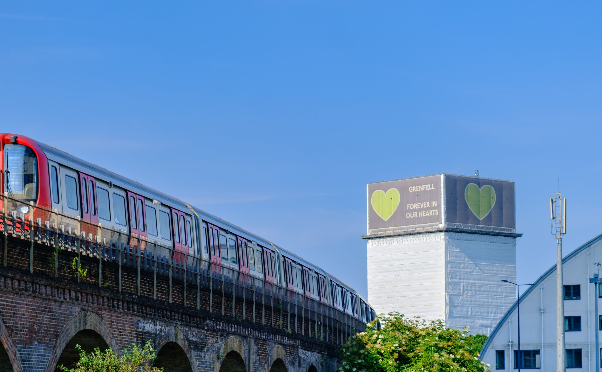 Grenfell Tower covered in white sheeting with a green heart banner reading “Grenfell – Forever in Our Hearts,”. A red London Underground train passes on a viaduct in the foreground.