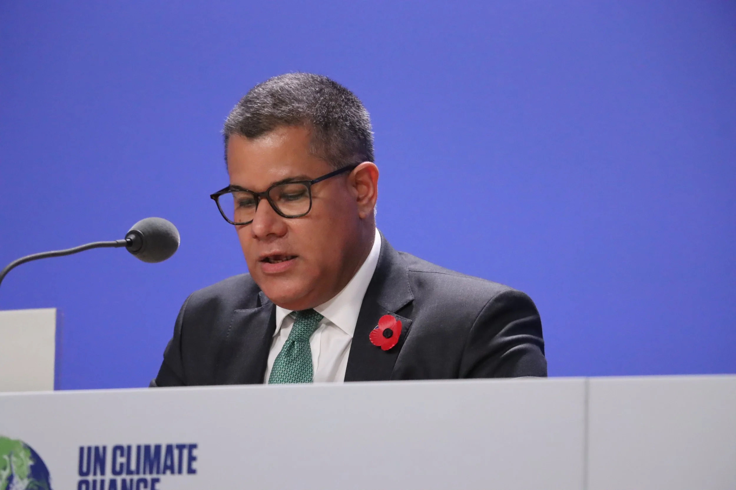 Lord Alok Sharma speaking at a podium during the COP26 climate summit, wearing a dark suit and poppy pin, with a "UN Climate Change" logo visible in front.
