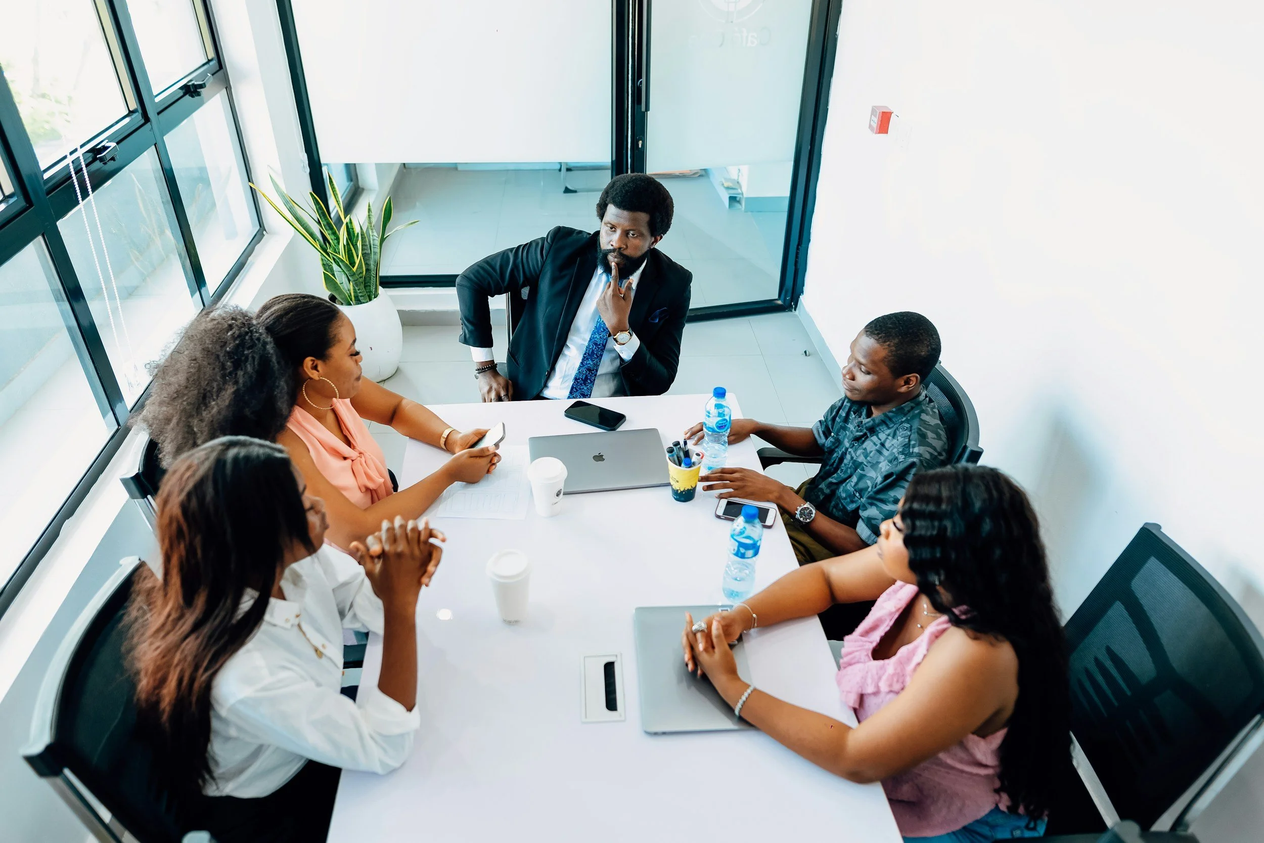 A group of six diverse professionals, including five women and one man, are having a meeting or discussion in a modern conference room with large windows, a potted plant, and a glass door.