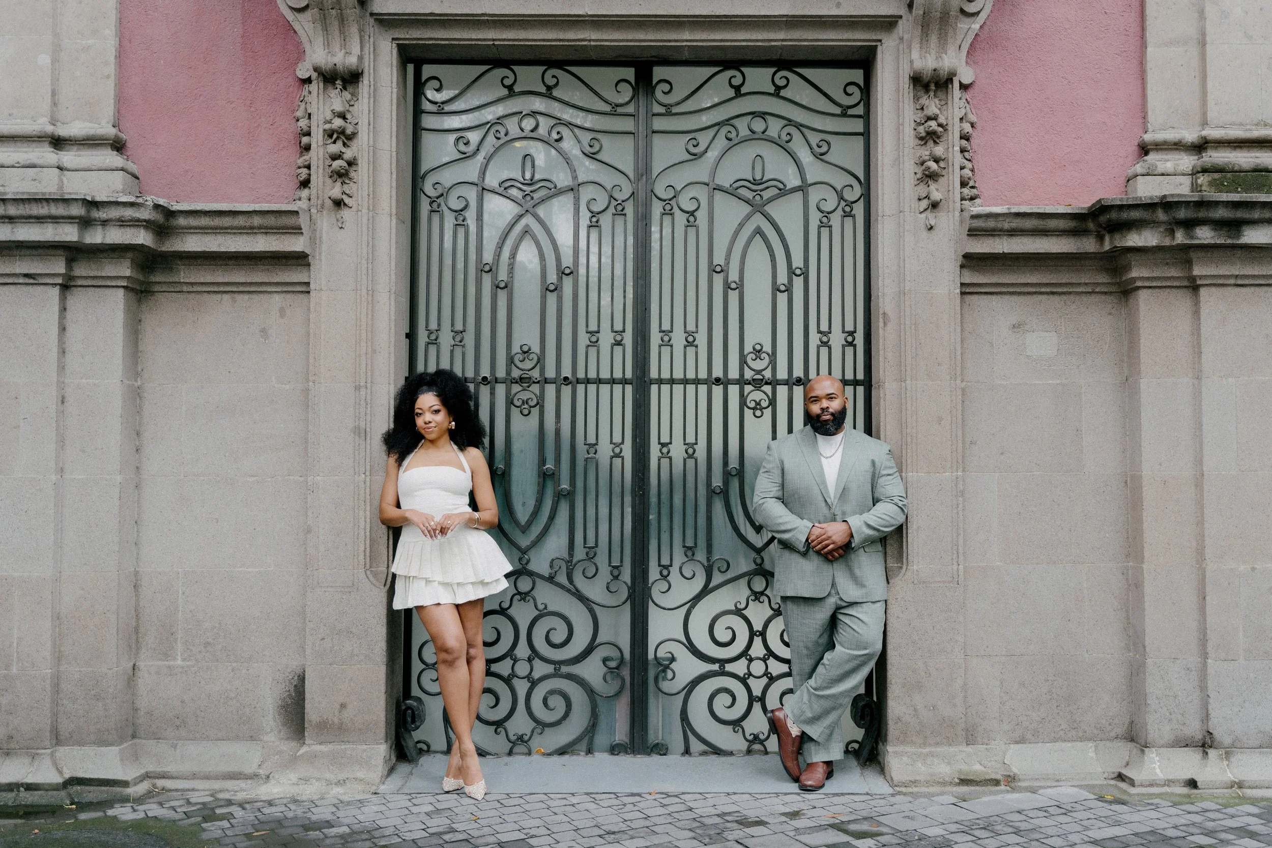 A woman in a white dress and a man in a gray suit with brown shoes standing against an ornate metal gate, in front of a pink and stone building.