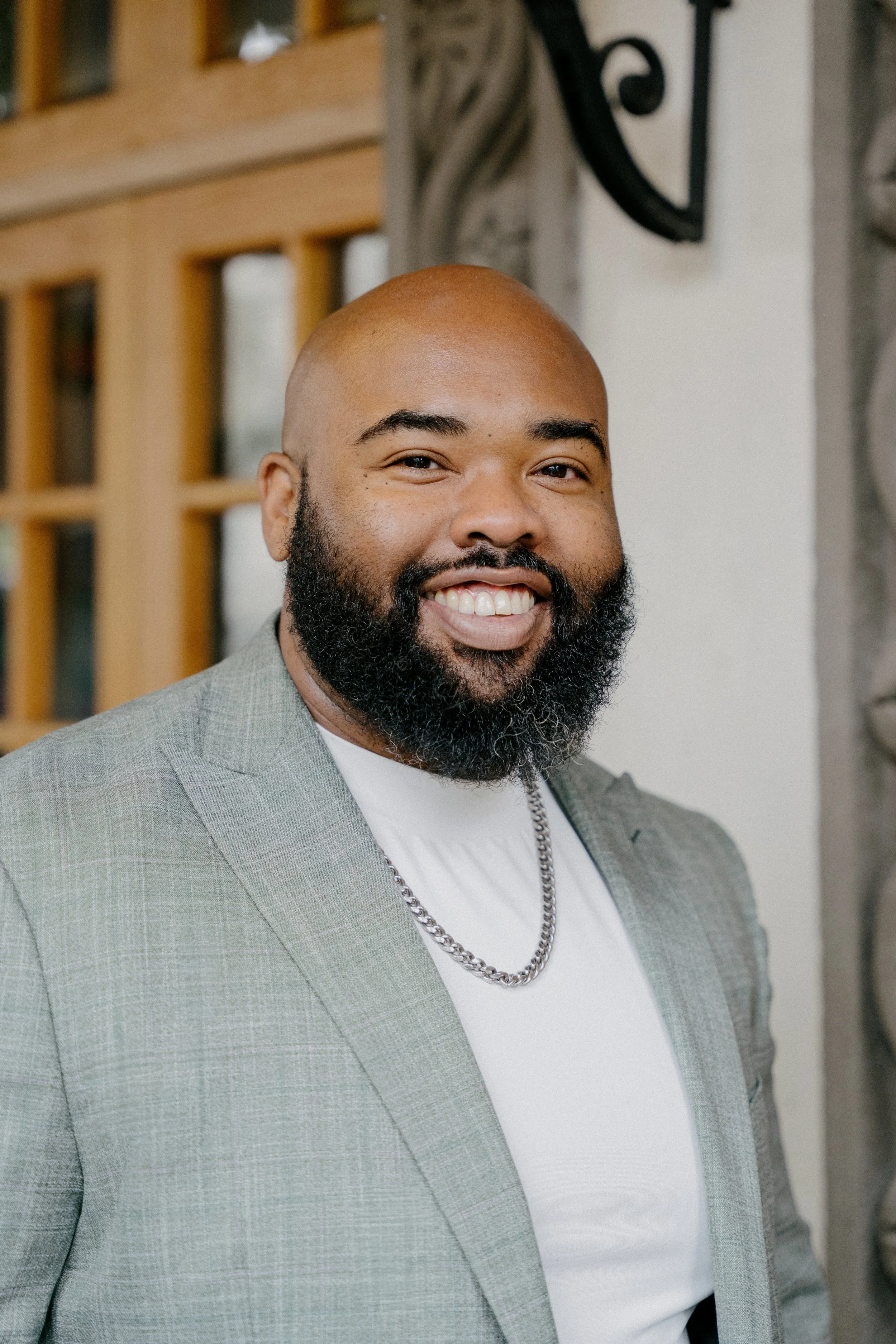 A smiling African American man with a beard and bald head, wearing a light gray plaid blazer, white t-shirt, and a silver chain necklace, standing in front of a wooden and stone building exterior.