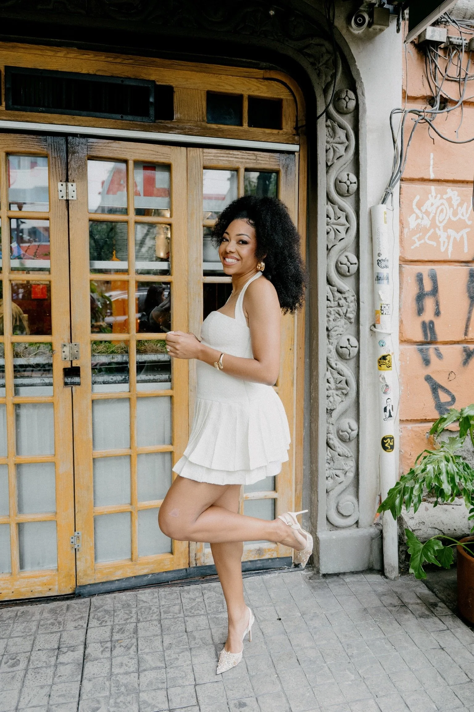 A woman with curly black hair wearing a white dress and high heels, standing on one leg in front of a wooden and glass door outside a building.