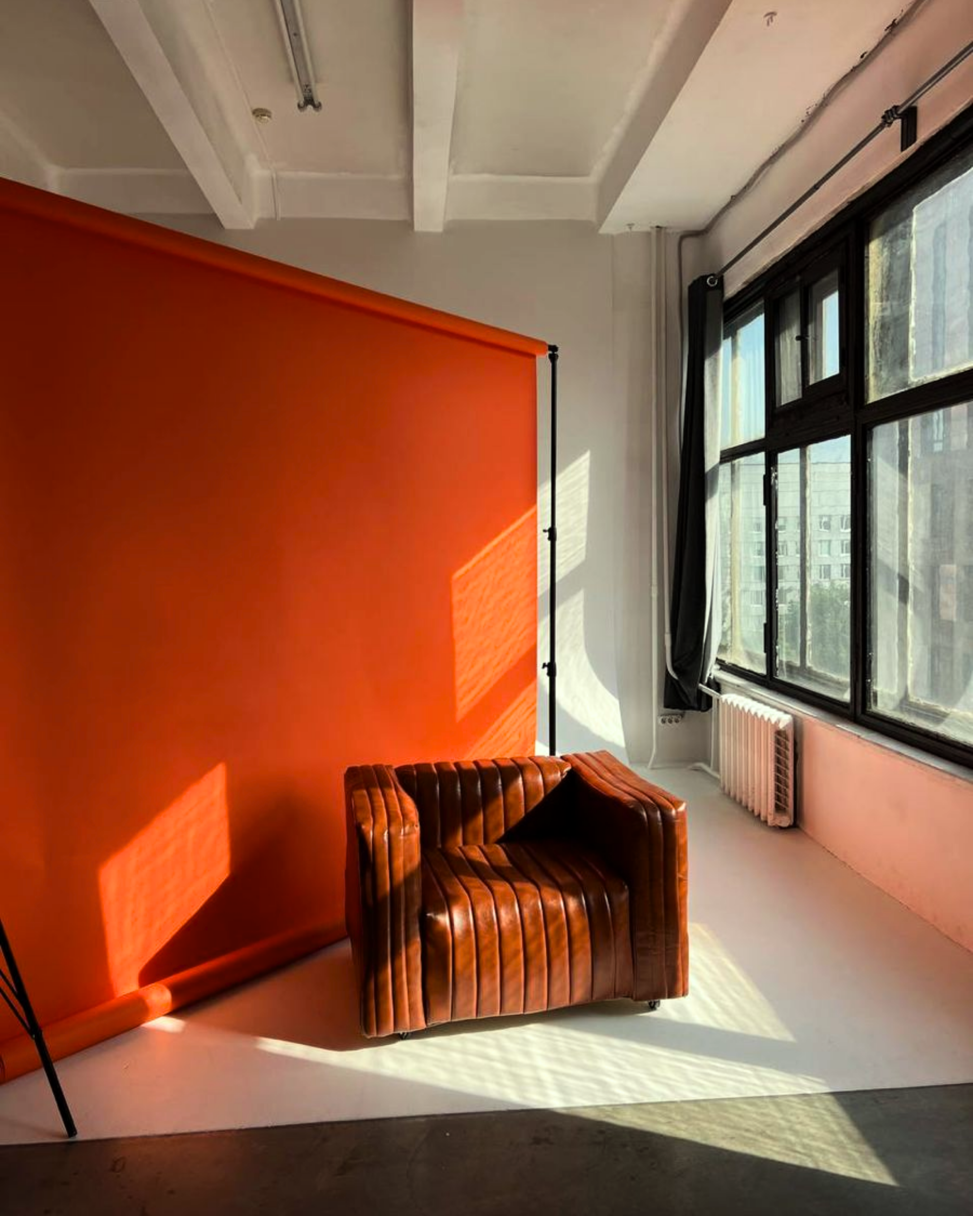 A brown leather armchair in a room with large windows, black curtains, and an orange backdrop, with sunlight casting shadows on the floor.