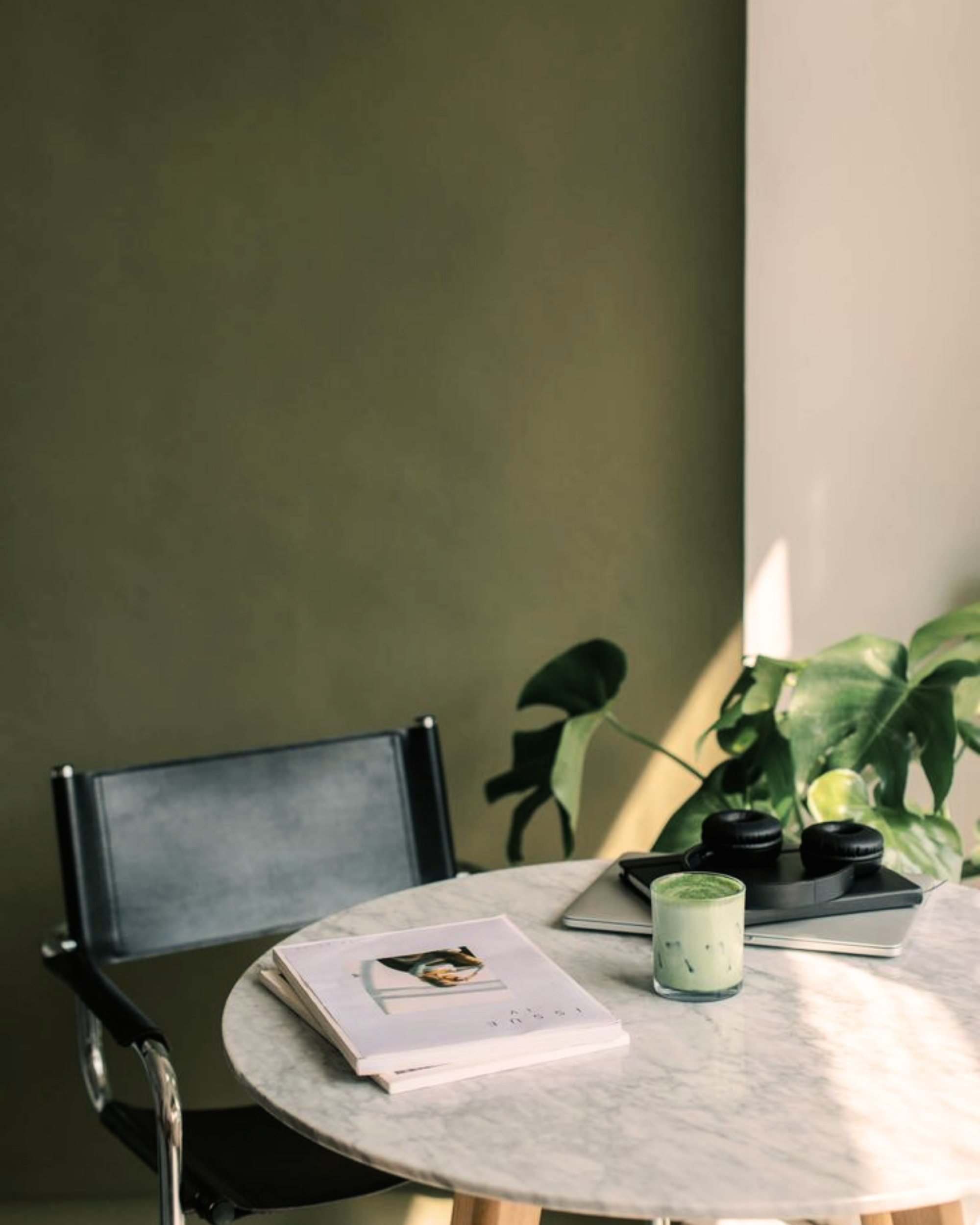 Round marble table with magazines, a laptop, headphones, and a green smoothie, surrounded by green plants and a black chair.