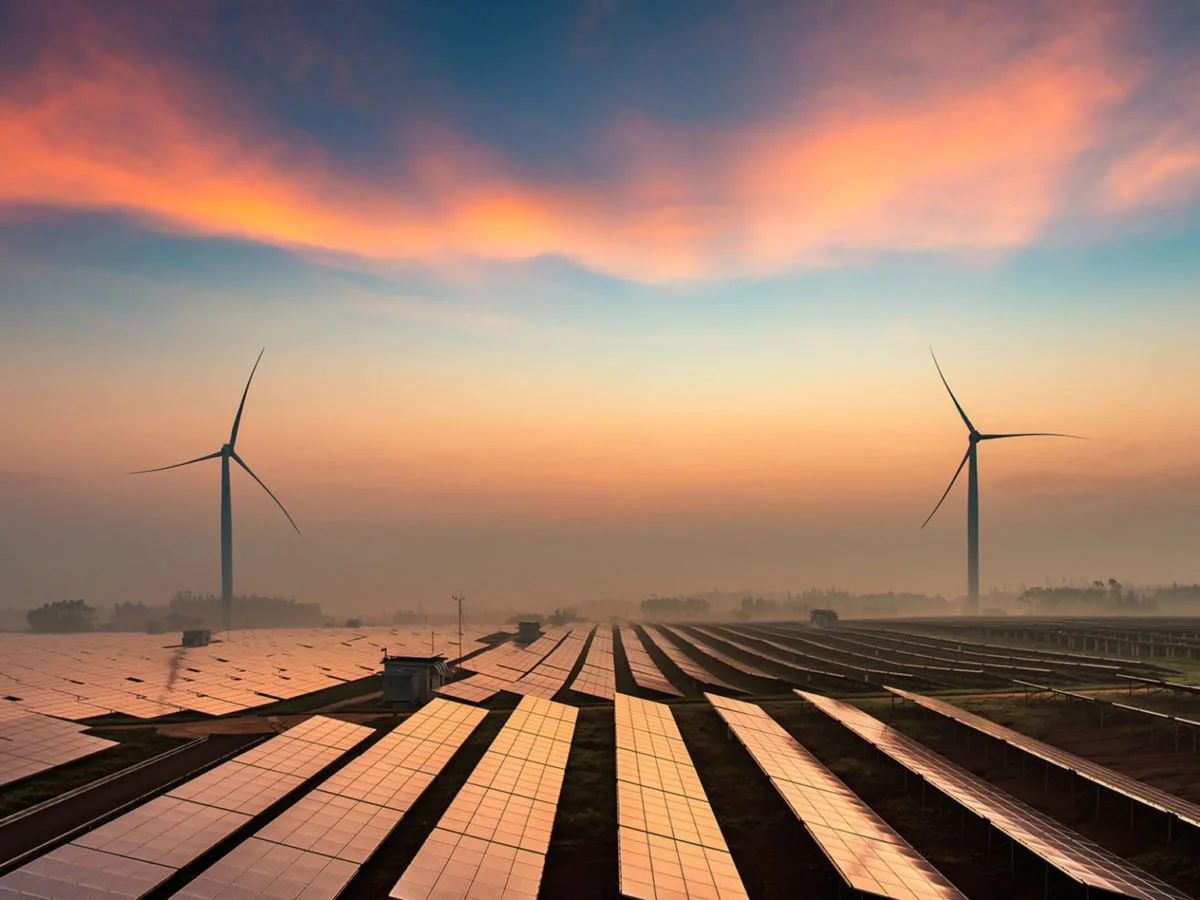 A solar farm with rows of solar panels in the foreground and wind turbines in the background during sunset.