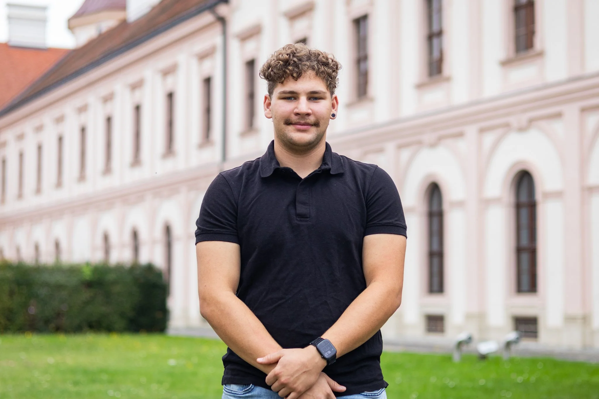 Junger Mann mit dunklem T-Shirt und Armbanduhr steht vor einem historischen Gebäude mit großen, arched Fenstern, grüner Wiese im Vordergrund.