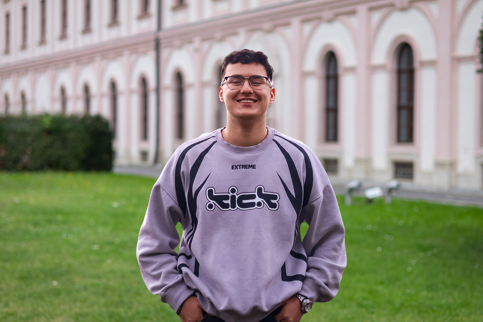 Ein junger Mann trägt ein graues Sweatshirt mit schwarzen Streifen und einem Logo, lächelt vor einem historischen Gebäude mit grüner Wiese im Hintergrund.