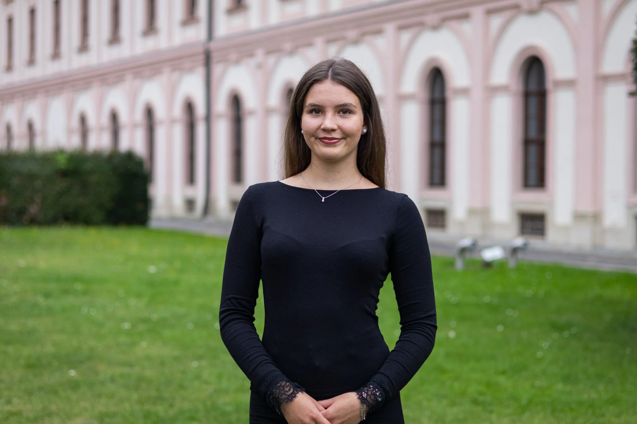 Eine junge Frau in schwarzem Kleid steht im Freien vor einem historischen pinken Gebäude mit grüner Wiese und Bänken im Hintergrund.