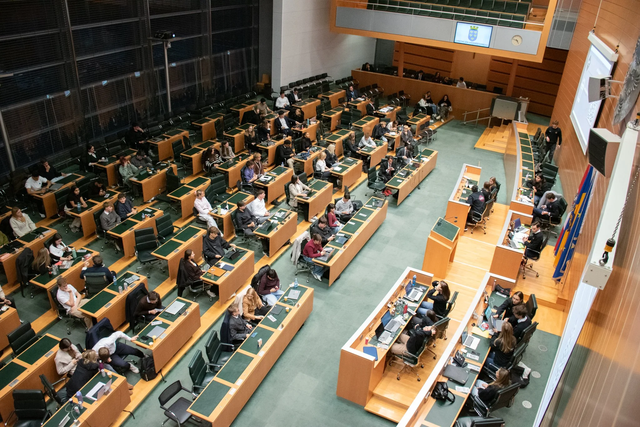 Verschiedene Menschen sitzen an Holztischen in einem großen, modernen Sitzungssaal. Einige arbeiten an Laptops, andere sprechen miteinander. Im Hintergrund ist eine große digitale Anzeige und Flaggen stehen an der Seite.
