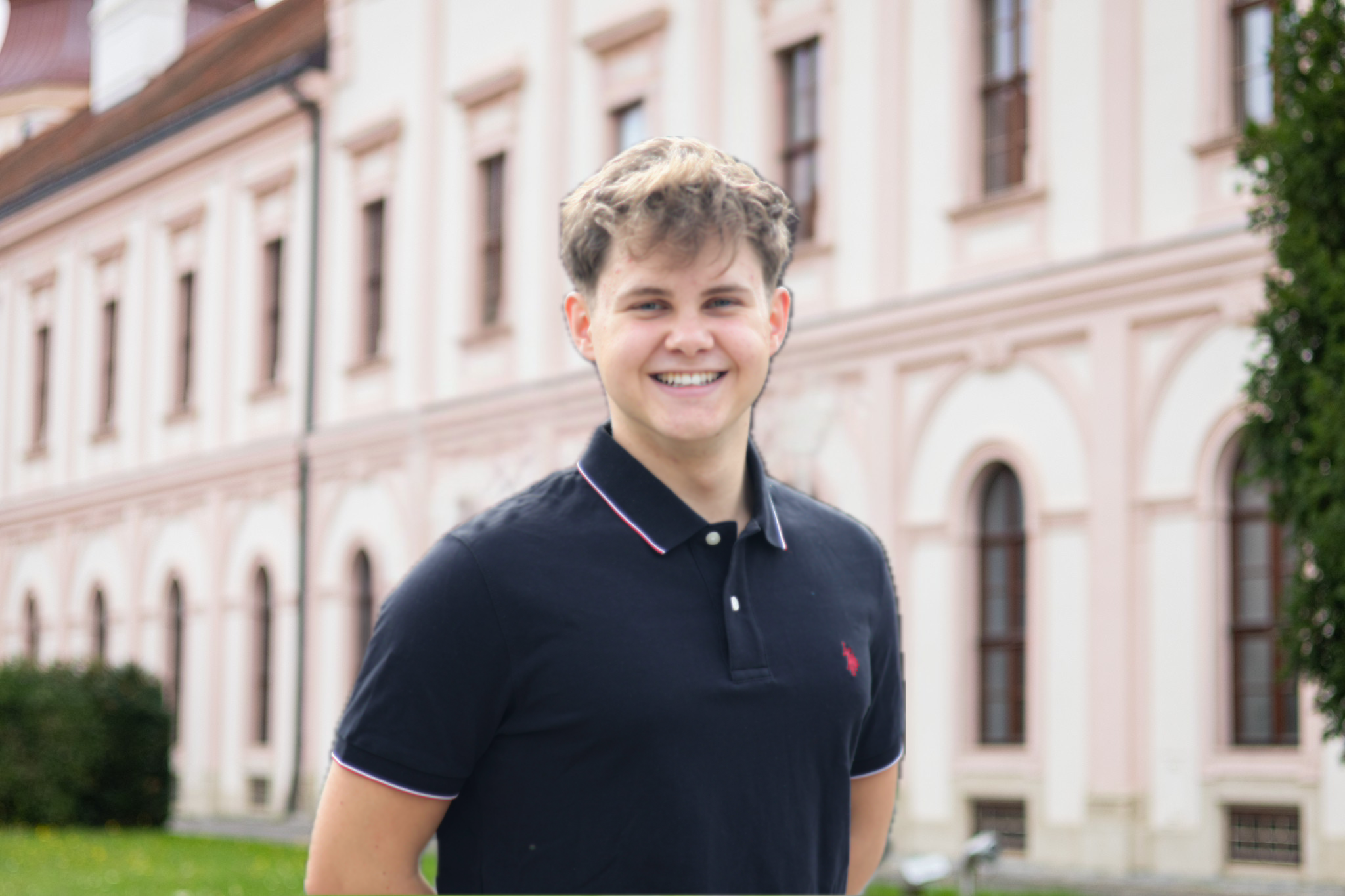 Ein junger Mann mit braunen, lockigen Haaren, lachend, trägt ein schwarzes Polo-Shirt, steht vor einem mehrstöckigen pinkfarbenen Gebäude mit großen Fenstern.