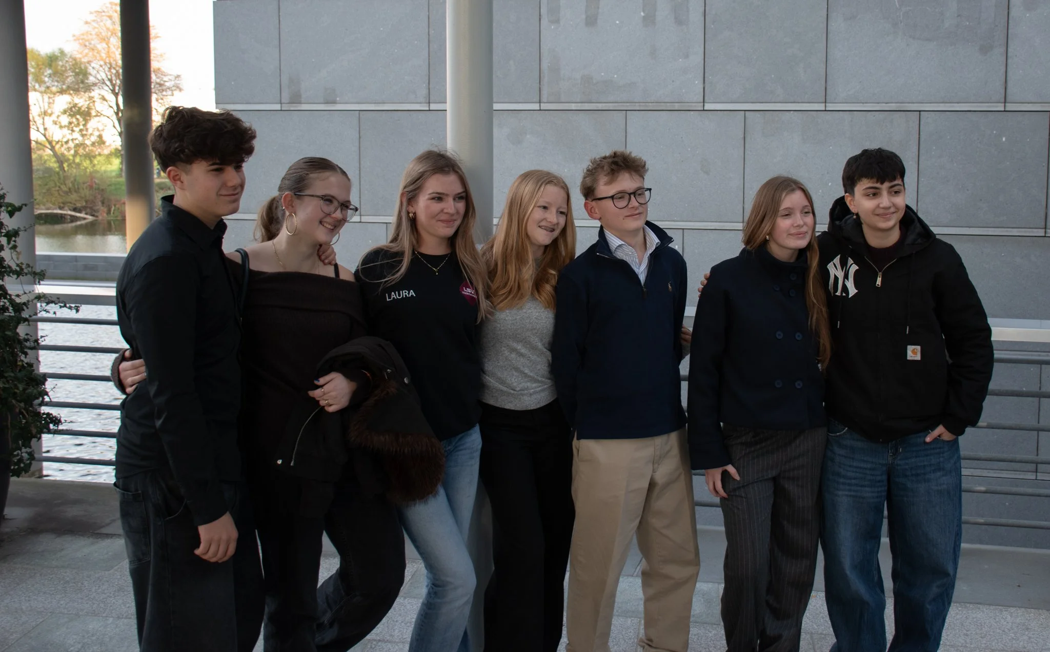 Gruppe von sieben jungen Menschen steht zusammen vor einer grauen Wand bei Sonnenuntergang.