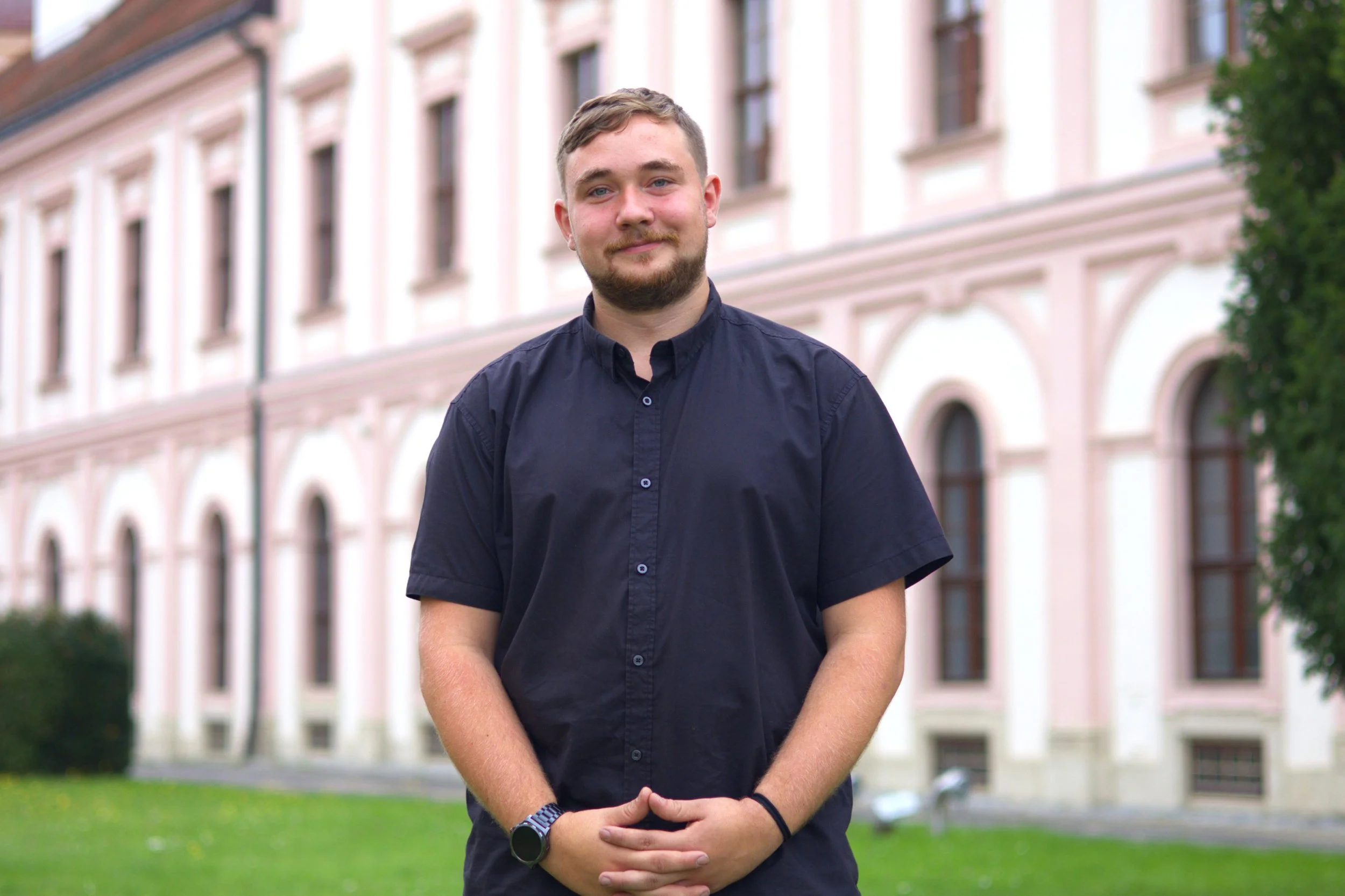 Junger Mann mit kurzen braunen Haaren und Bart trägt ein dunkelblaues Hemd, steht vor einem pinkfarbenen Gebäude mit vielen Fenstern auf einer grünen Wiese.
