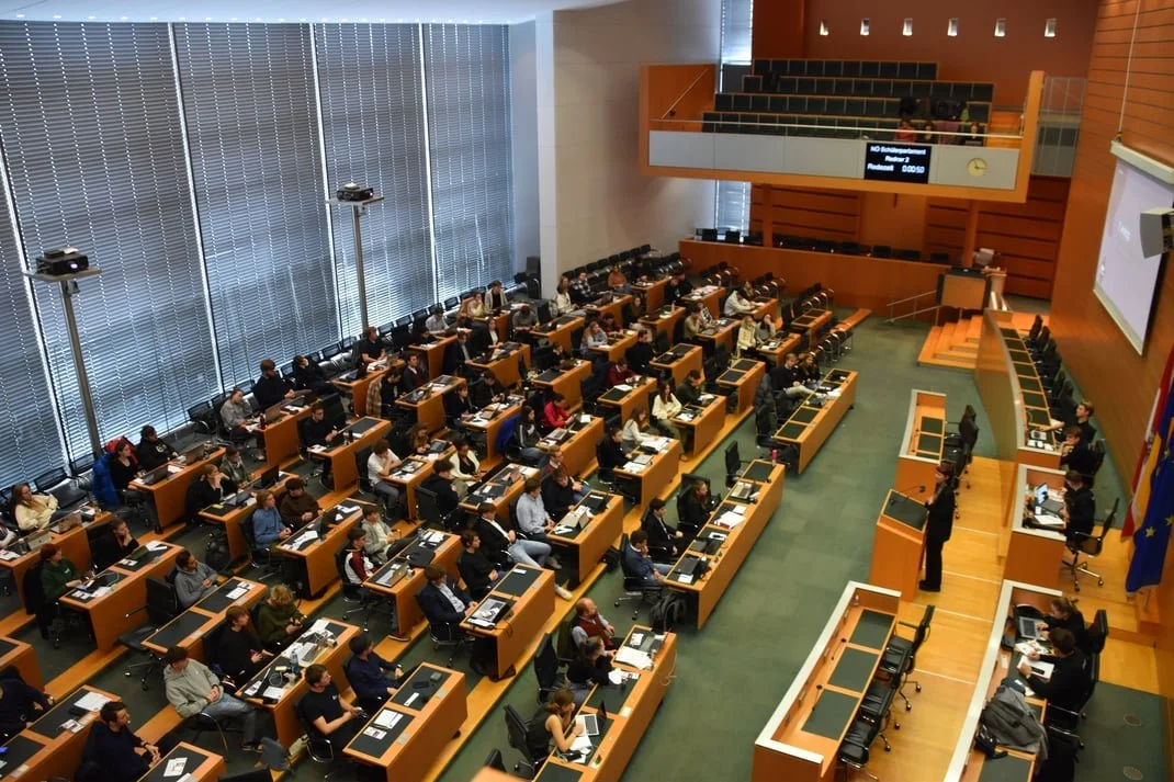 Verschiedene Menschen sitzen in einem großen Sitzungssaal mit Holzsitzen, auf einem erhöhten Podium befindet sich eine Person, die spricht, im Raum sind deutsche und europäische Flaggen, der Raum hat große Fenster mit vertikalen Jalousien.