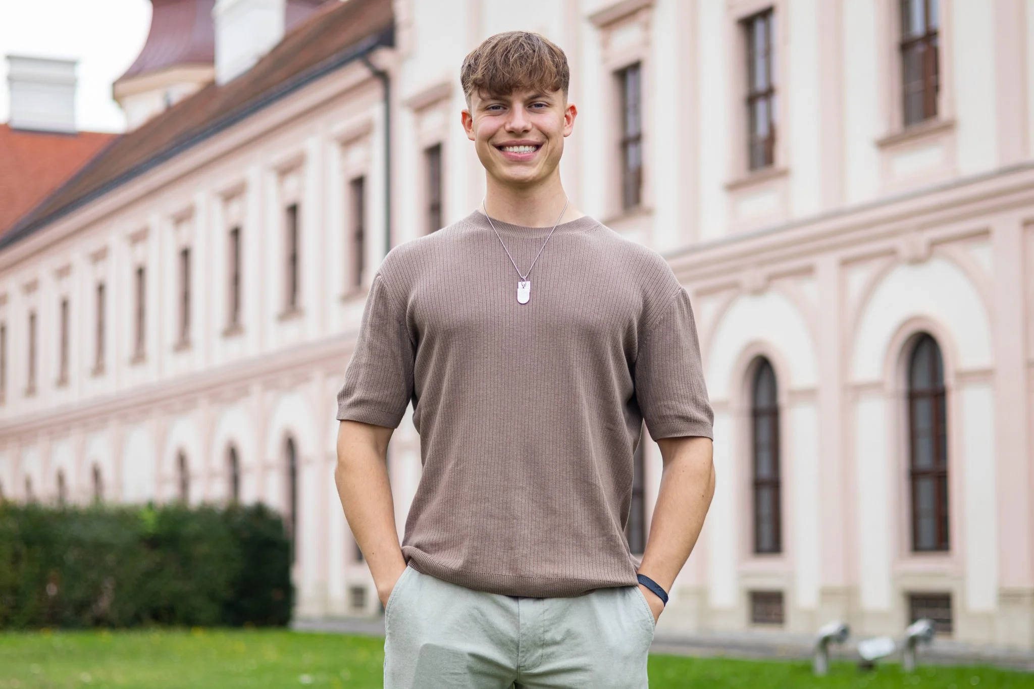 Ein junger Mann in beigem T-Shirt und hellen Hosen steht vor einem historischen Gebäude mit großen Fenstern und rotem Dach, lächelnd in die Kamera schauend.