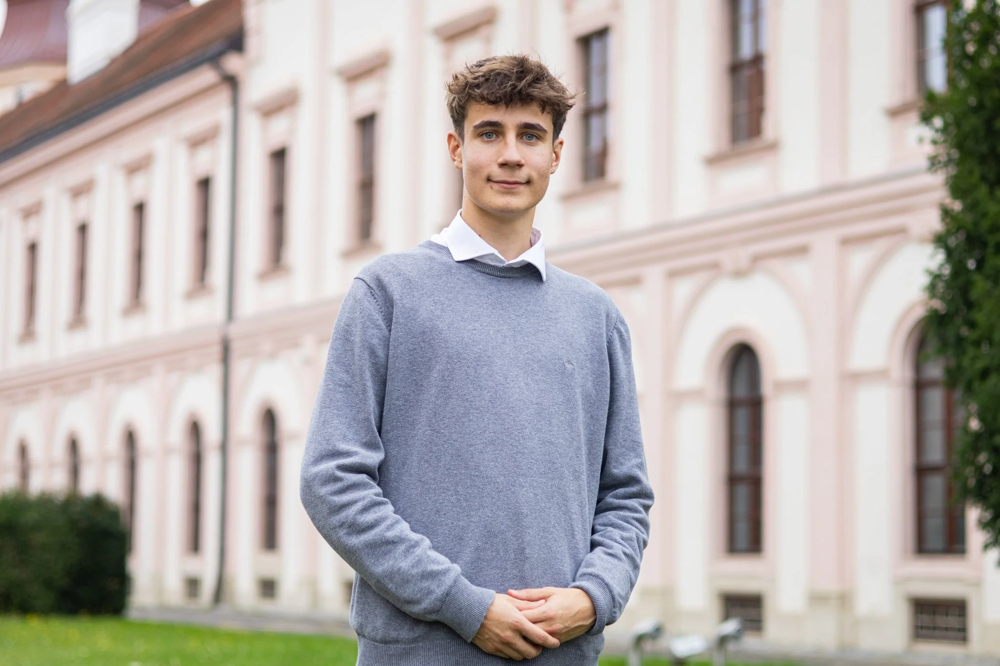 Ein junger Mann steht vor einem historischen Gebäude mit großen Fenstern und einem grünen Garten im Hintergrund.