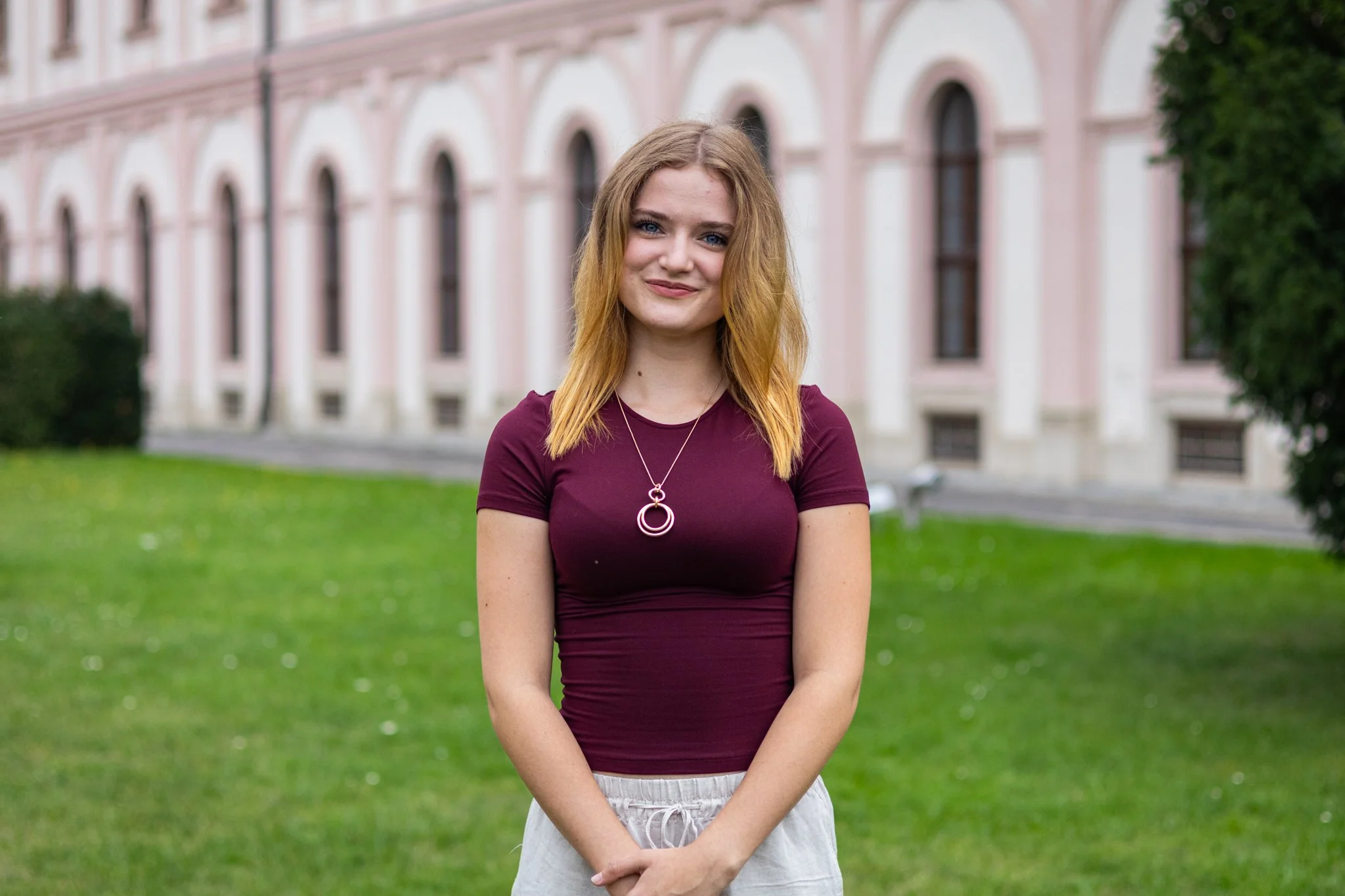 Junge Frau mit blonden Haaren, blauer Augen und einem burgunderfarbenen T-Shirt steht vor einem grünen Rasen und einem historischen Gebäude mit weißen Säulen, im Freien.
