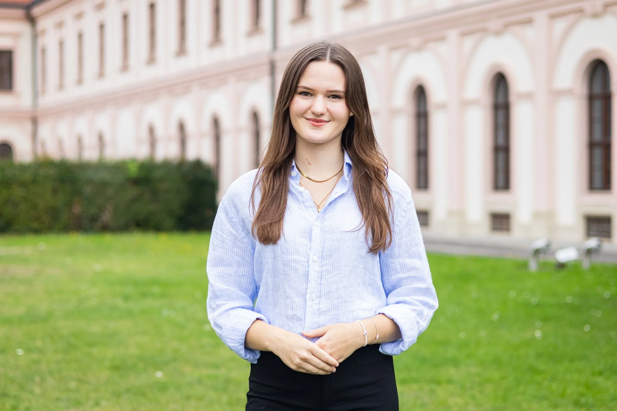Junge Frau mit braunen Haaren und blauer Hemdbluse, die im Freien vor einem historischen Gebäude steht.