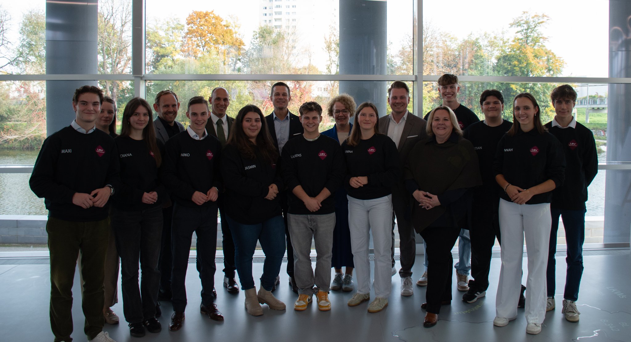 Gruppe von jungen Menschen mit Erwachsenen in einem modernen Gebäude mit großen Fenstern und Blick auf Bäume im Herbst.