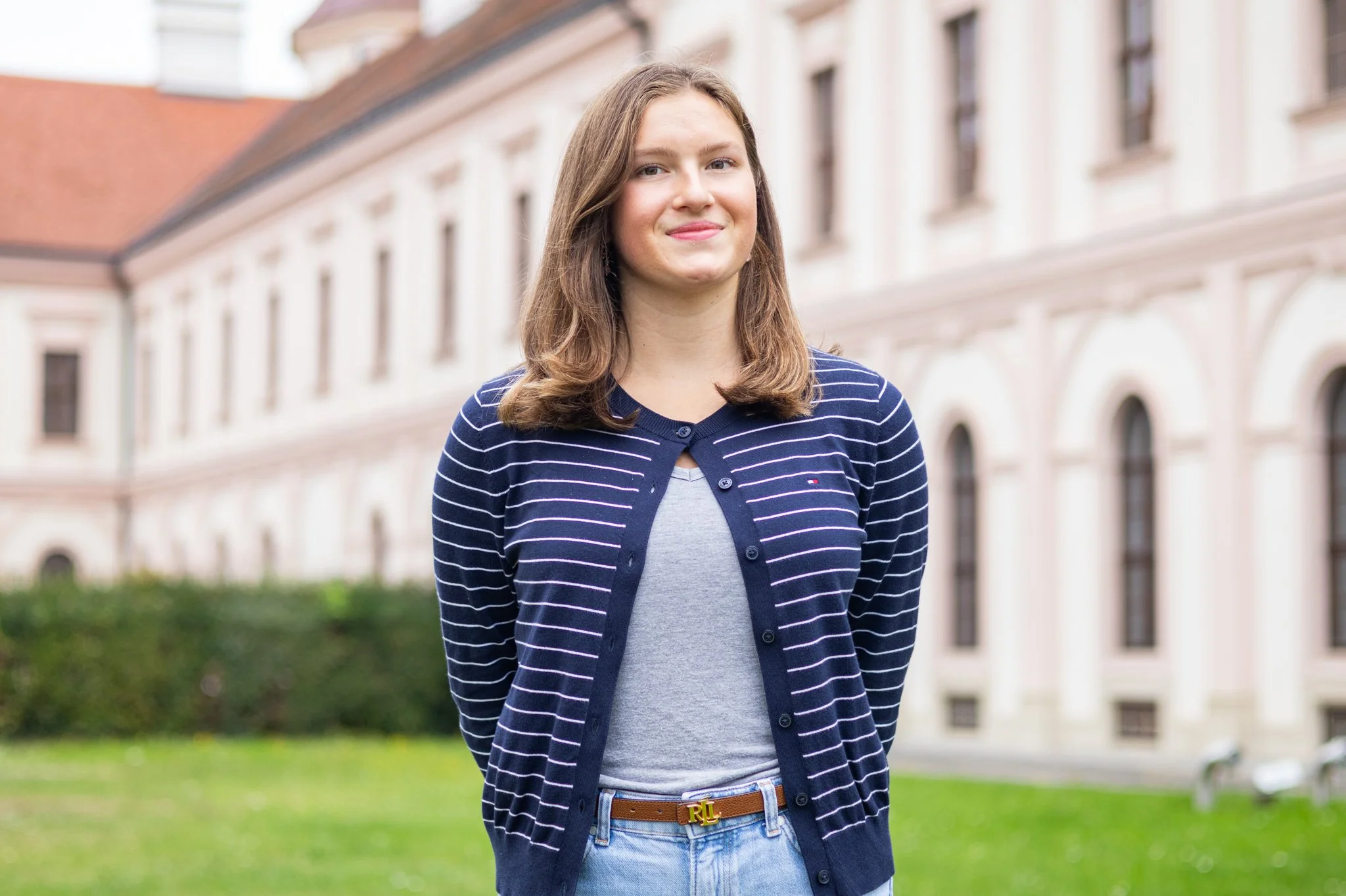 Junge Frau steht im Freien vor einem historischen Gebäude mit einer Gartenanlage im Vordergrund, trägt ein gestreiftes dunkelblaues Cardigan, ein graues T-Shirt, eine braune Gürtel und lächelt leicht.