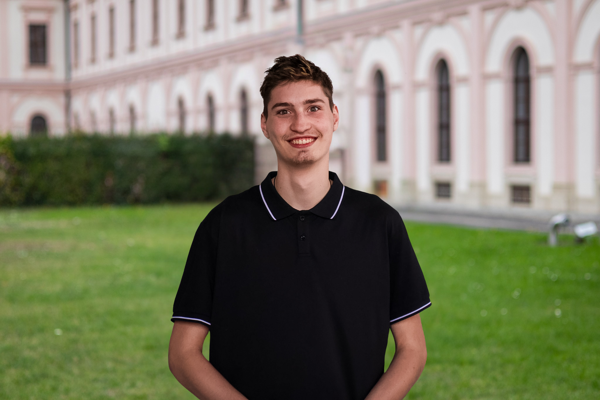 Ein junger Mann mit schwarzen Polo-Shirt steht vor einem historischen Gebäude mit roten und weißen Wänden und großen Fenstern, auf einer grünen Rasenfläche.