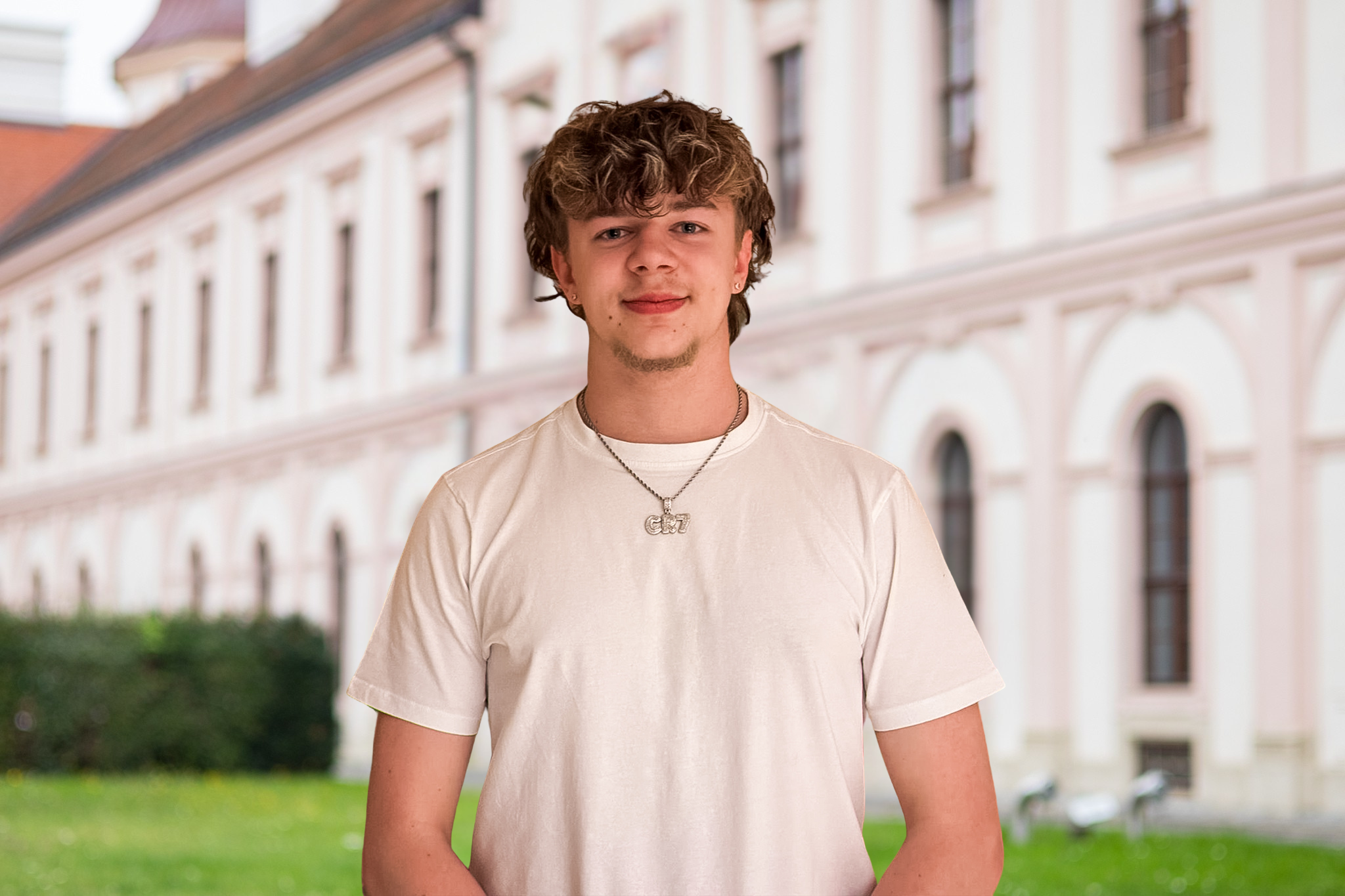 Junger Mann trägt ein weißes T-Shirt und eine Halskette mit einem Anhänger 'GZT' vor einem historischen Gebäude mit weißen Säulen und grüner Wiese.
