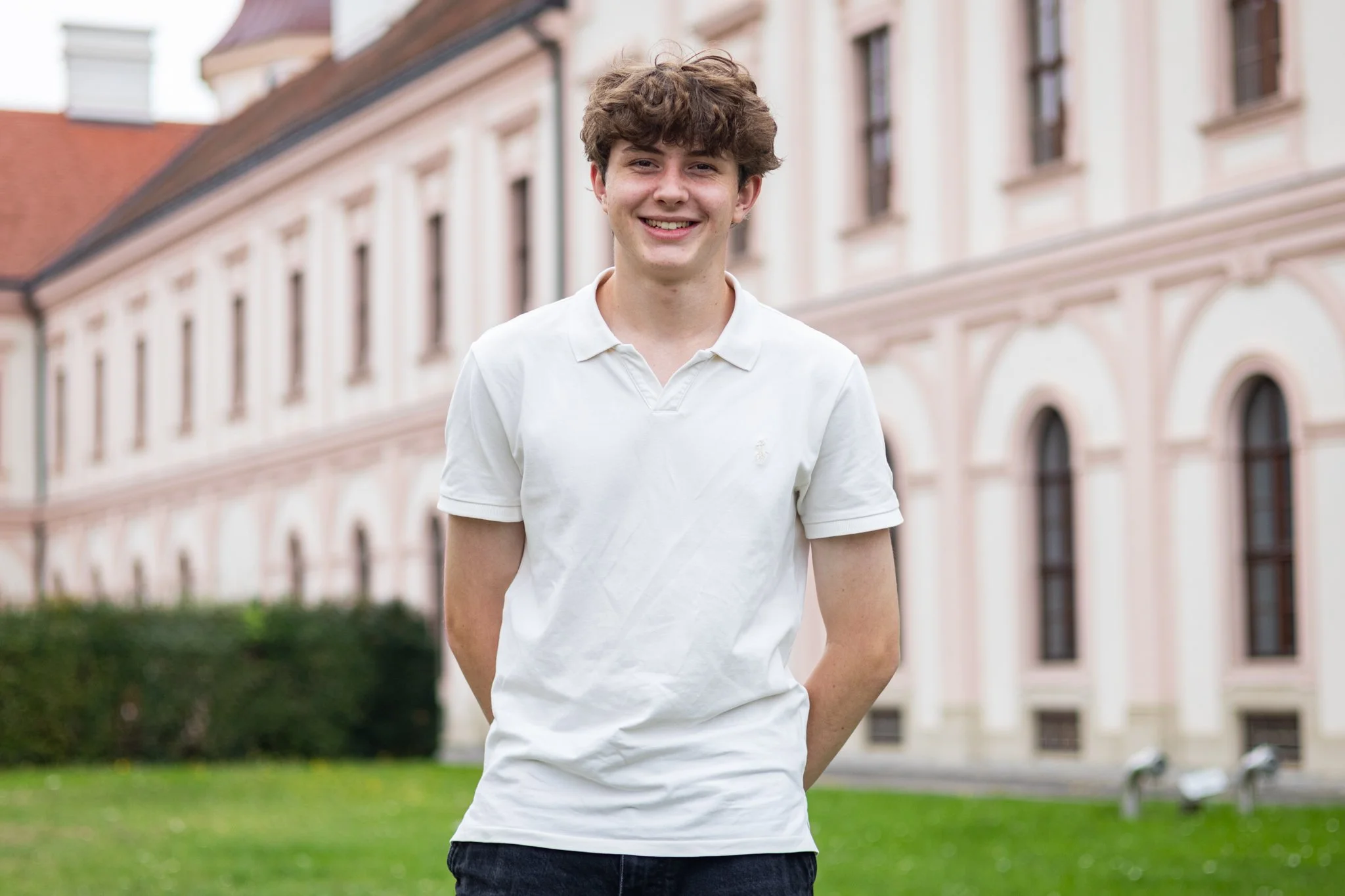 Ein junger Mann mit braunen, lockigen Haaren, trägt ein weißes Poloshirt, steht draußen auf einer Wiese vor einem historischen Gebäude mit großen Fenstern und roten Ziegeldächern, lächelt in die Kamera.