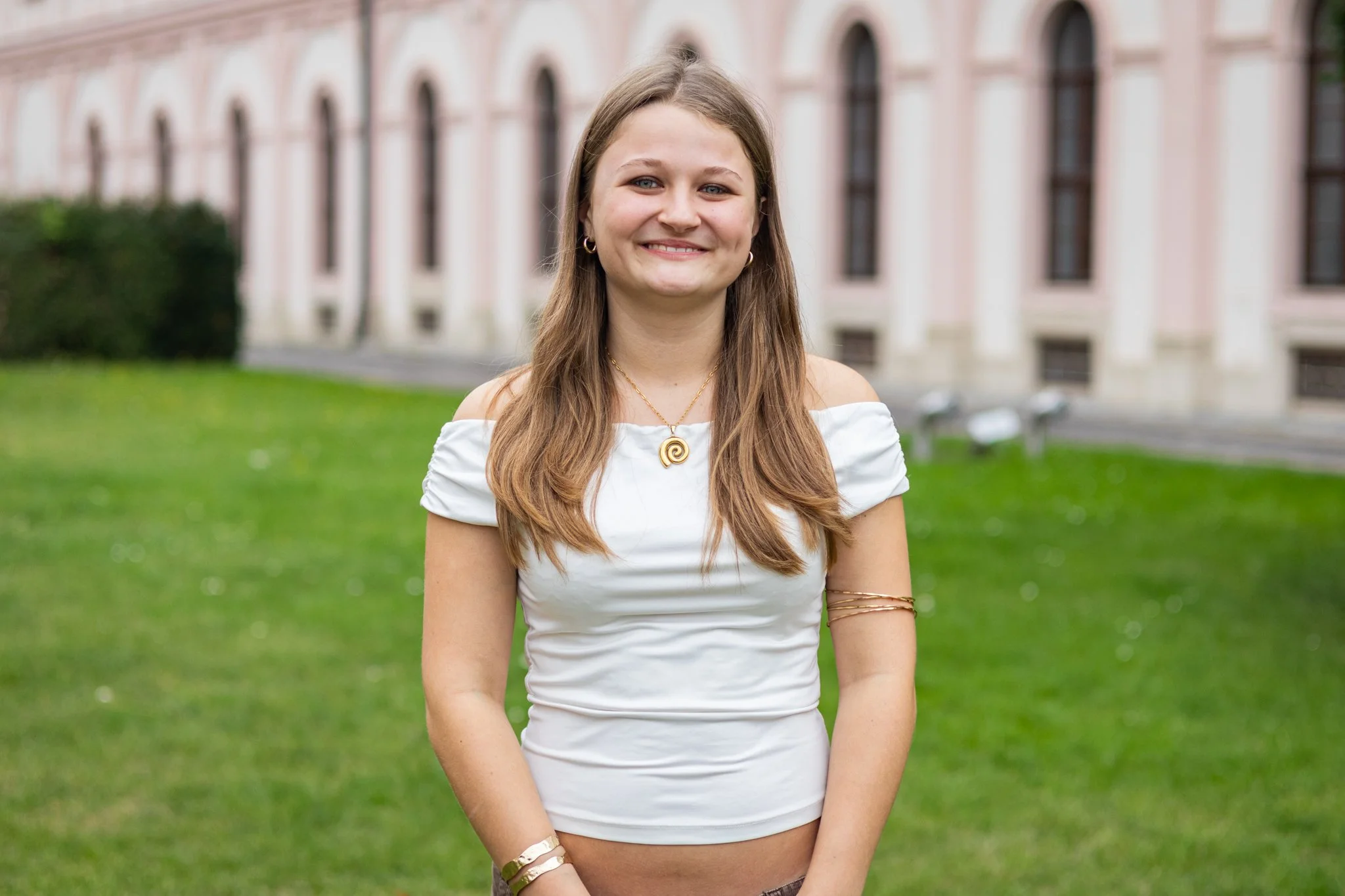 Junge Frau mit braunen, langen Haaren, weißem Oberteil und goldenen Schmuck, stehend auf einer grünen Wiese vor einem historischen Gebäude, lächelnd.