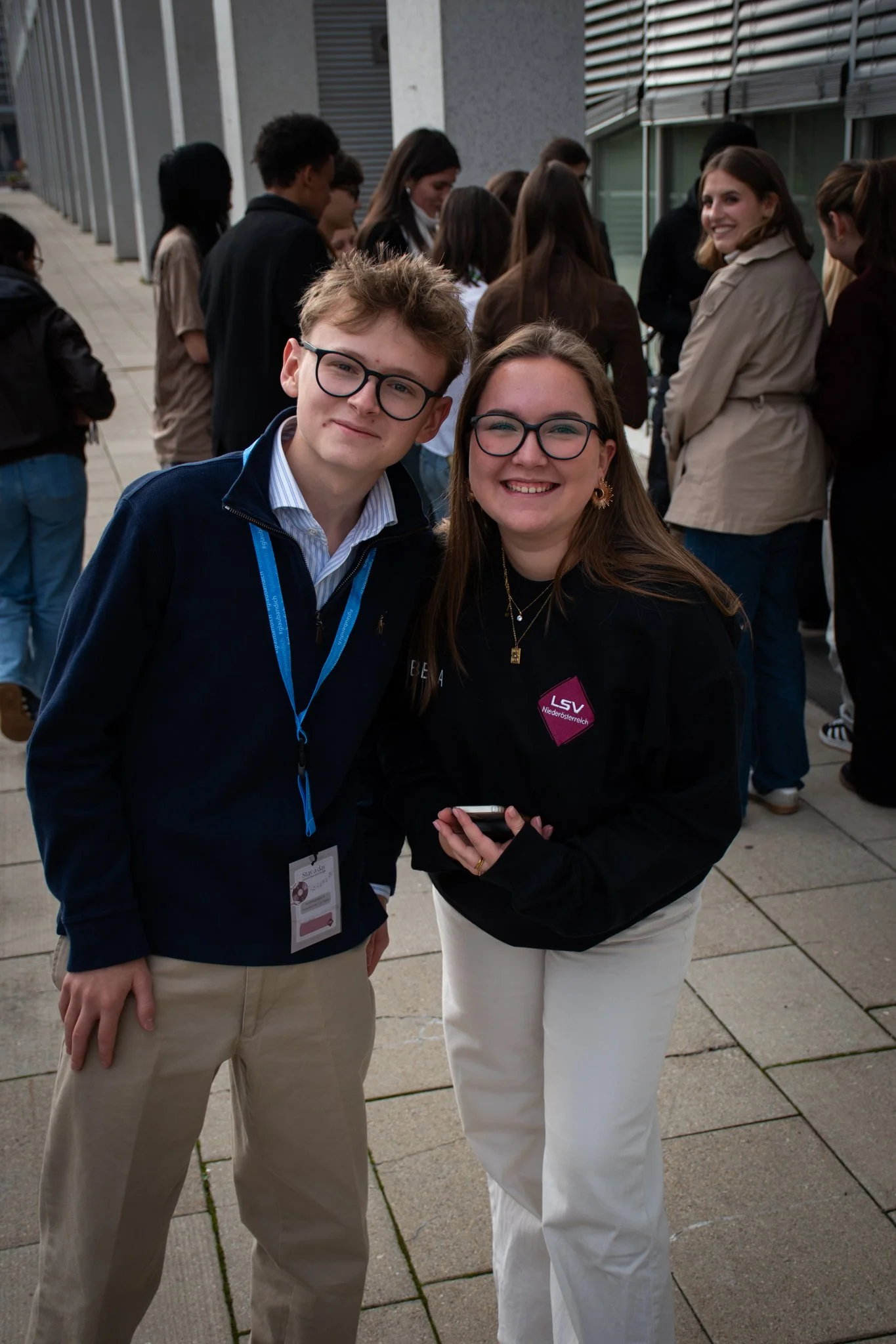 Zwei junge Menschen, ein Mann und eine Frau, lächeln in die Kamera bei einer Veranstaltung im Freien, im Hintergrund stehen andere Personen.
