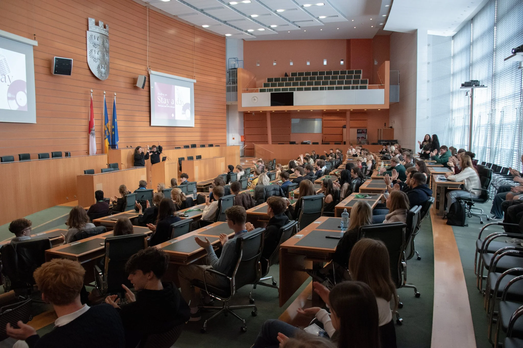 Ein großer Konferenzraum mit vielen Schülern, die an Tischen sitzen, und zwei Personen, die vorne auf einer Bühne sprechen. Es gibt große Leinwände mit Präsentationen, Flaggen an der Wand und große Fenster, die viel natürliches Licht einlassen.
