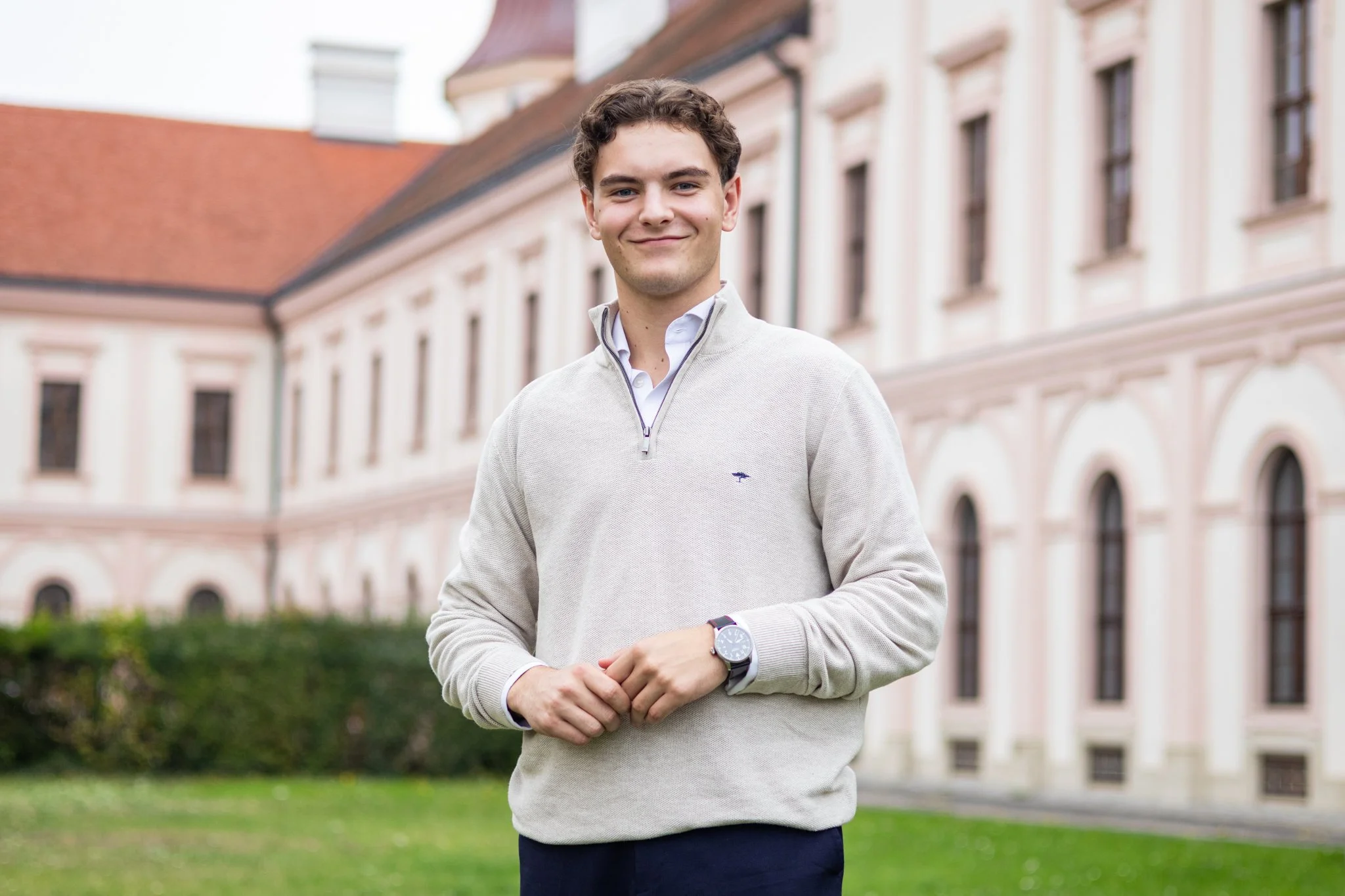Ein junger Mann steht vor einem historischen Gebäude mit rotem Dach und großen Fenstern, trägt einen hellgrauen Pullover und eine Uhr am Handgelenk, lächelt.