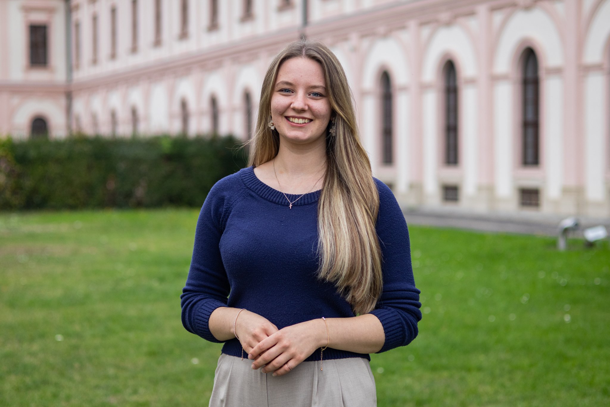 Junge Frau mit langem Haar, blaue Pullover, hellbeige Hose, im Freien vor einem historischen Gebäude mit weißen Säulen und großen Fenstern, lächelnd.