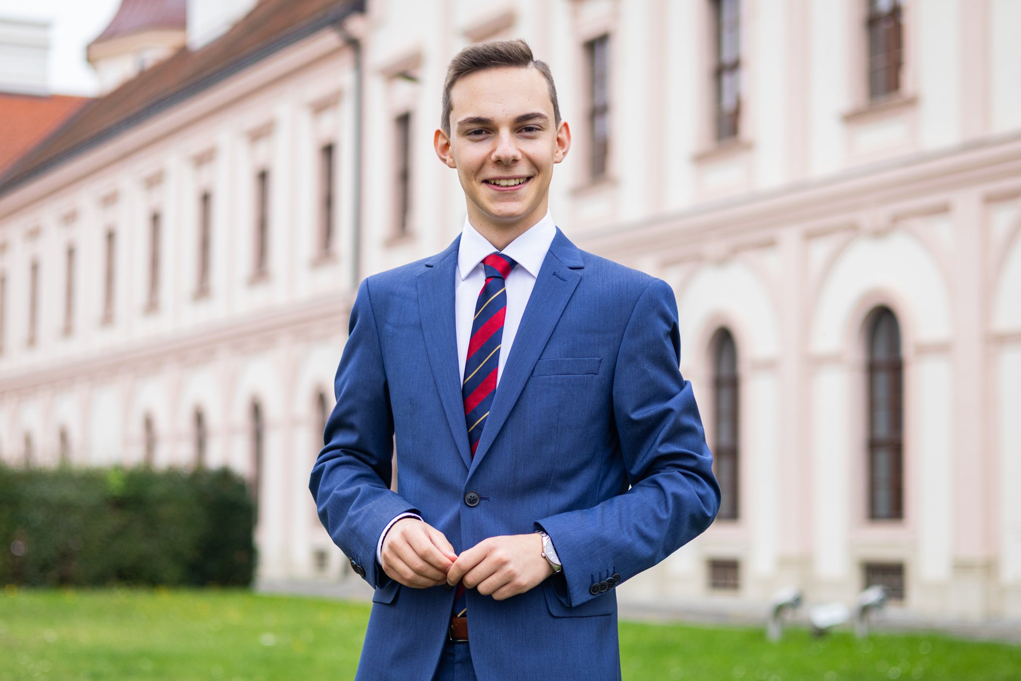 Ein junger Mann in einem blauen Anzug, weißem Hemd und einer diagonalen roten und blauen Krawatte steht vor einem historischen Gebäude mit mehreren Fenstern und einem roten Dach. Er lächelt und hat die Hände vor sich.