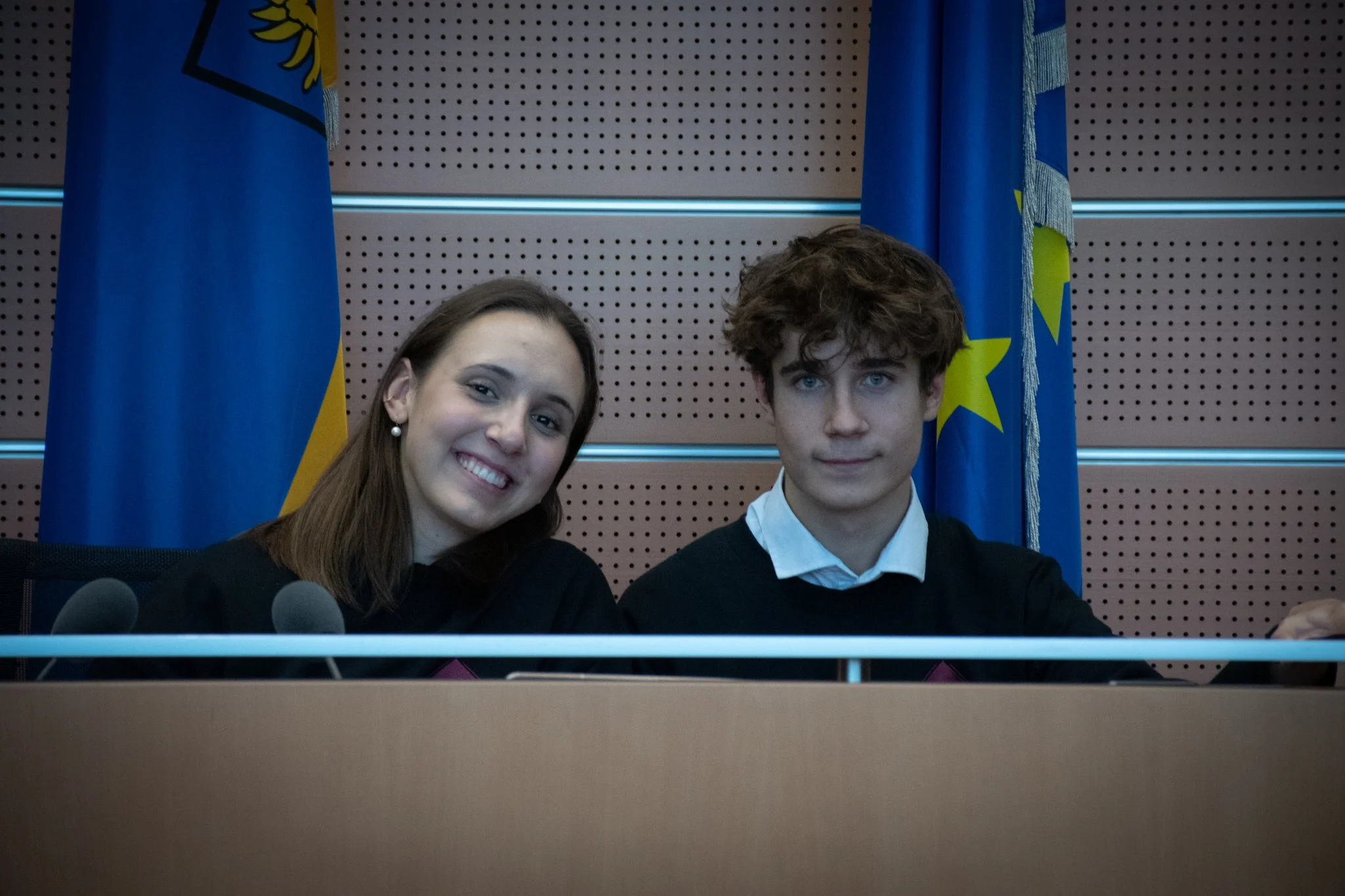 Zwei junge Menschen sitzen an einem Konferenztisch, mit europäischen und deutschen Flaggen im Hintergrund.