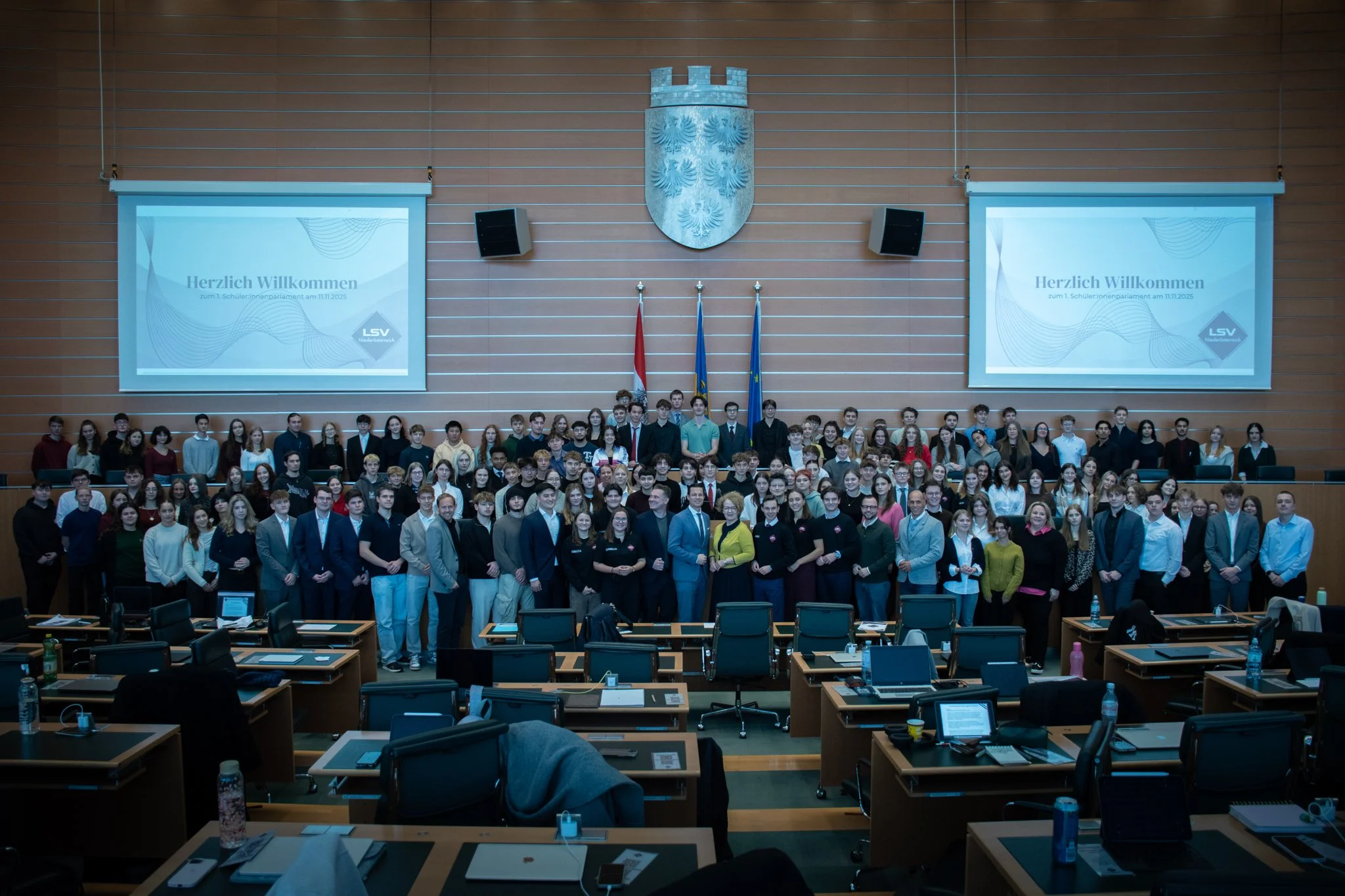 Gruppenfoto in einem großen Sitzungsraum mit vielen Menschen, die für eine Veranstaltung posieren. Im Hintergrund befinden sich zwei Bildschirme mit Willkommensnachrichten, Flaggen und ein Wappen an der Wand.