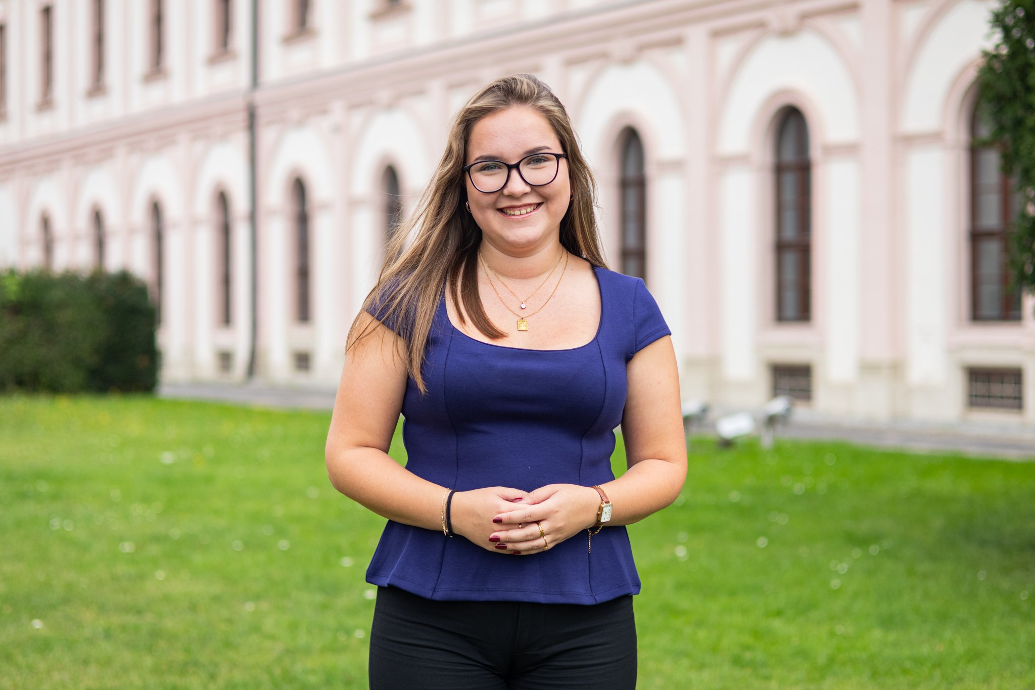 Lächelnde junge Frau in blauer Bluse mit Brille steht auf einem Rasen vor einem historischen Gebäude mit großen Fenstern.