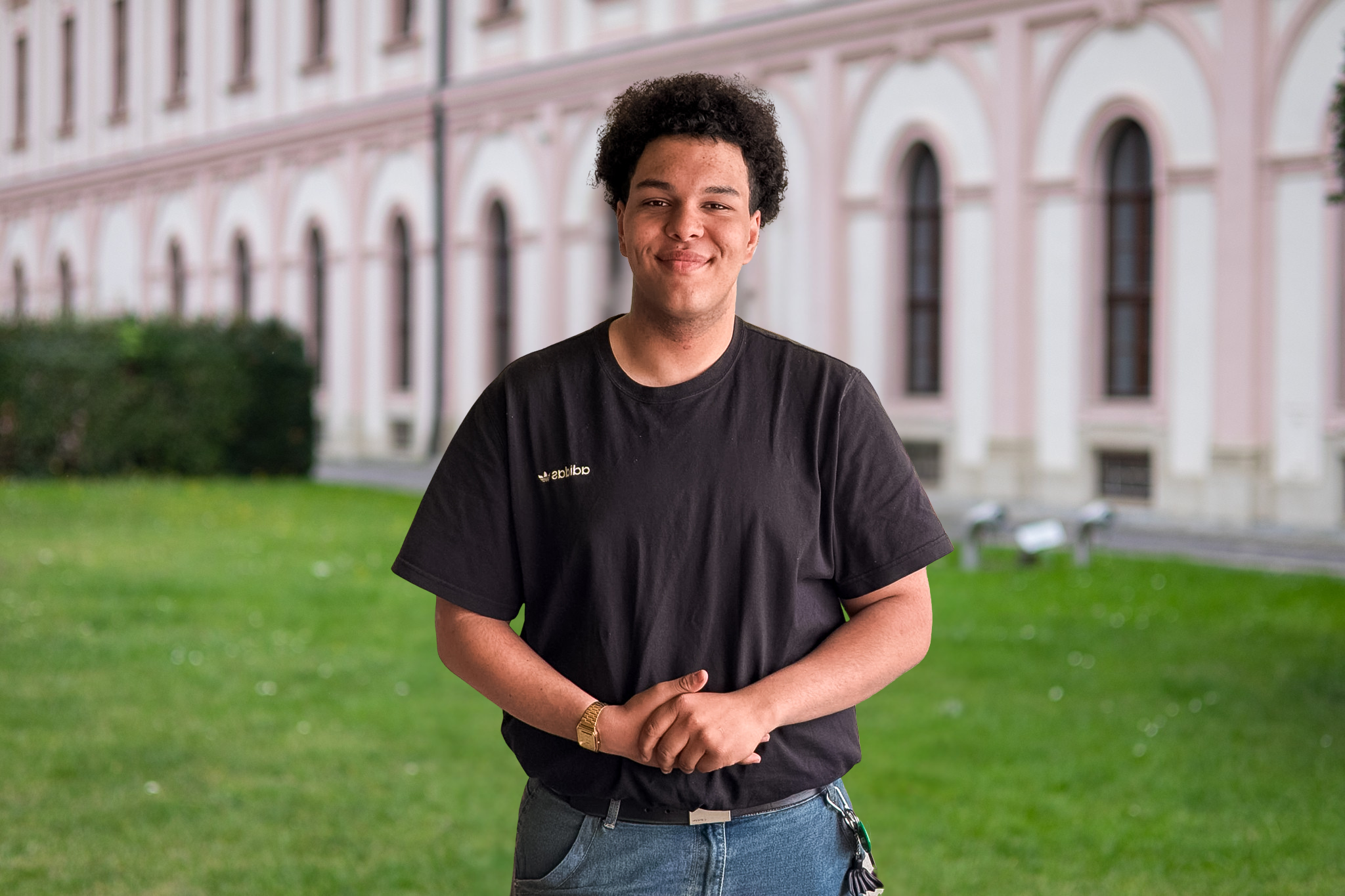 Junge Mann steht im Freien vor einem grünen Rasen und einem historischen Gebäude mit weißen Bögen, trägt schwarze T-Shirt, Jeans, Armbanduhr und Schlüsselbund.
