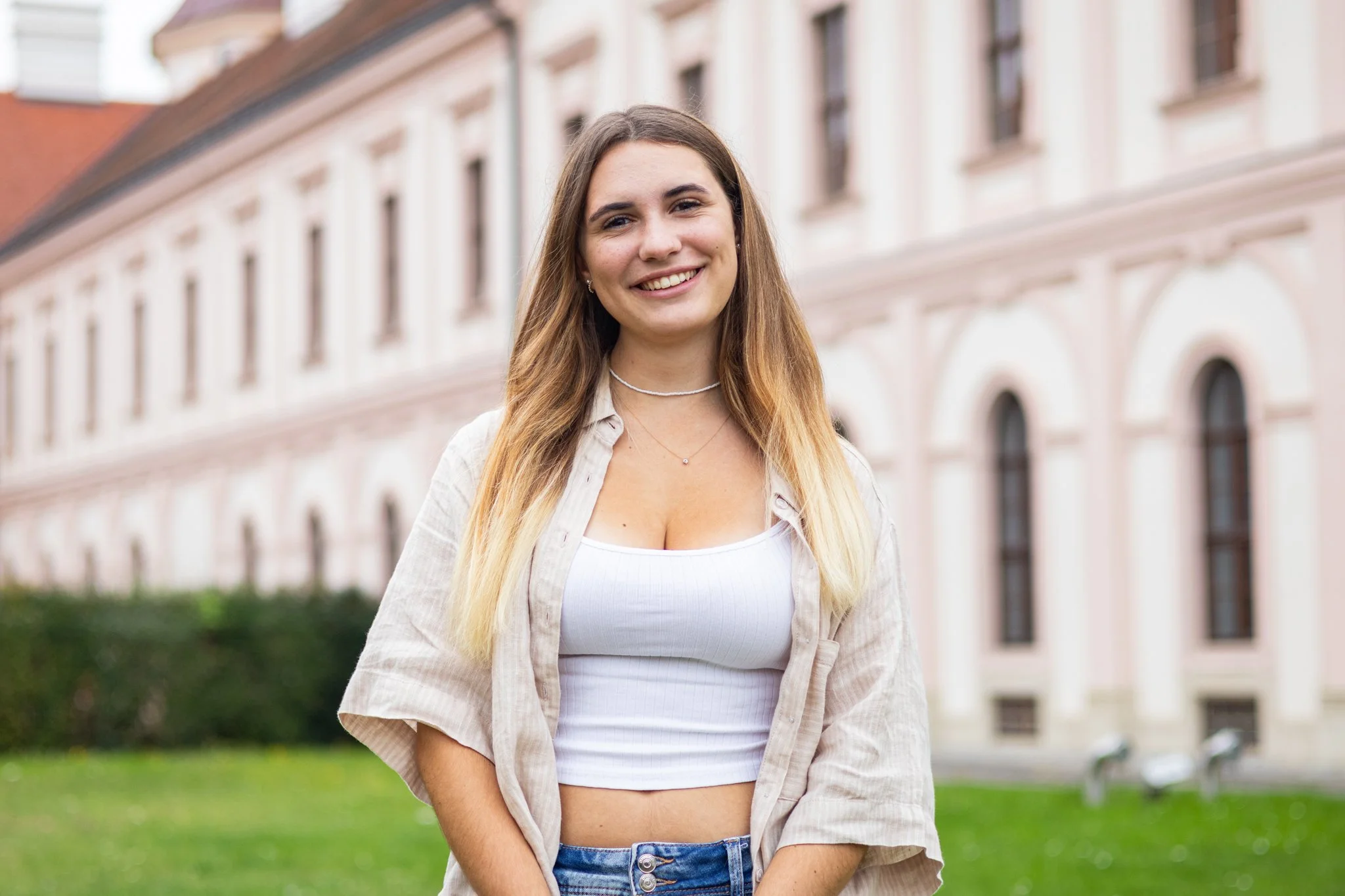 Eine lächelnde junge Frau mit langen, blond-braunen Haaren steht im Freien vor einem klassischen, mehrstöckigen Gebäude mit vielen Fenstern und einer rosa Fassade. Sie trägt ein weißes Crop-Top, eine offene Beige-Jacke, Jeans und Schmuck.