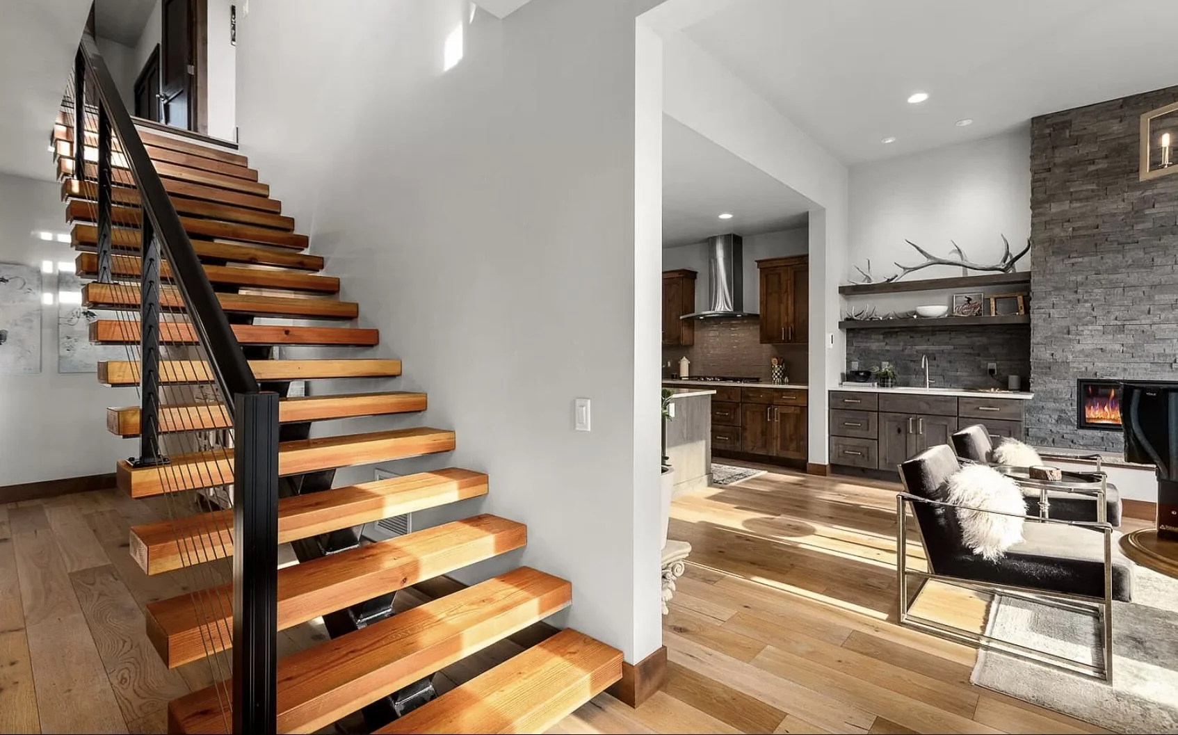 Interior of a modern home featuring a wooden staircase with black metal railing, hardwood floors, a living room area with leather and furry armchairs, a stone fireplace, and a kitchen with wooden cabinets and stainless steel appliances.